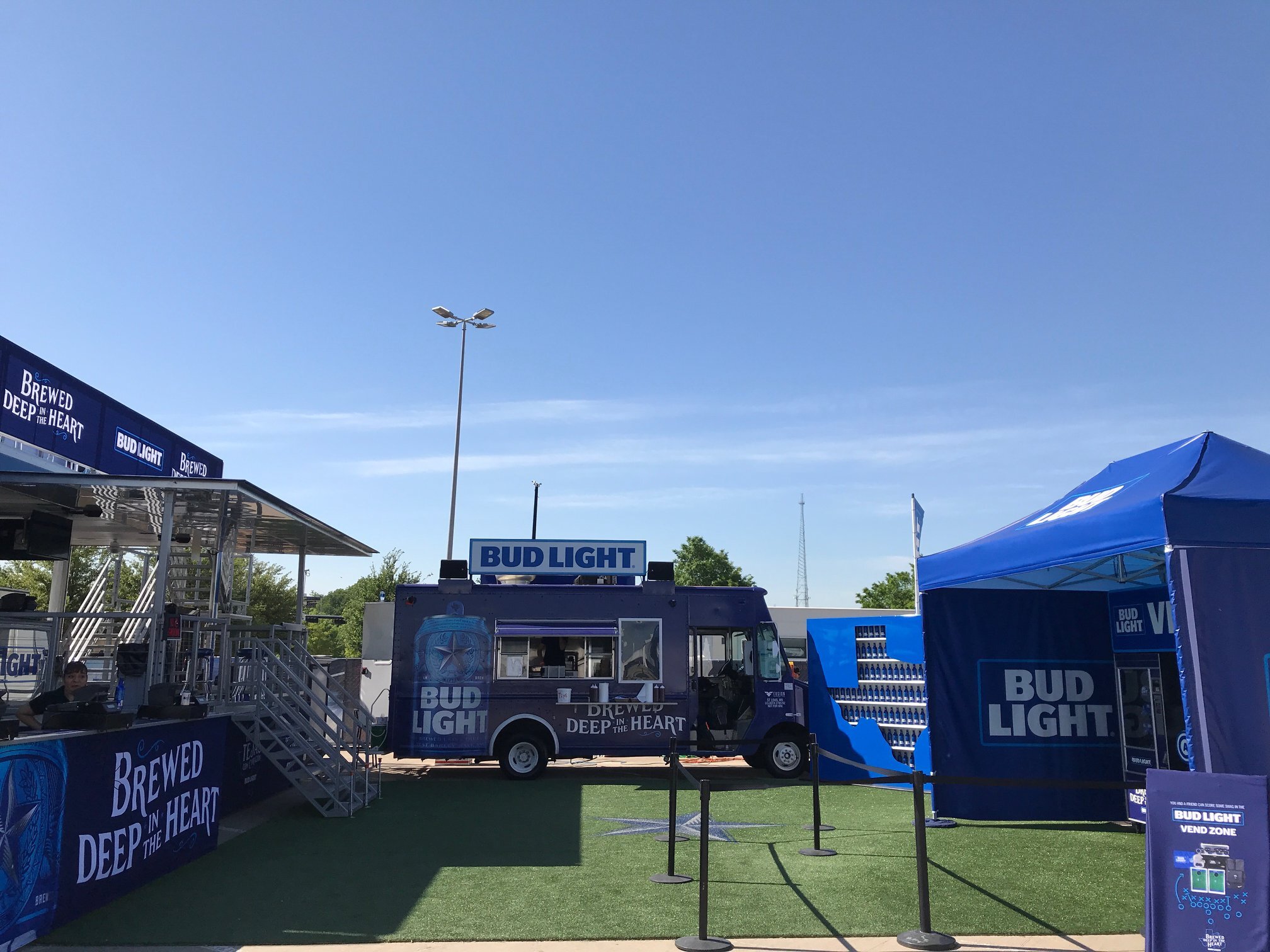 Outdoor area with BUD LIGHT branding, blue tents, and a vending trailer, set up for an event or festival on a sunny day.