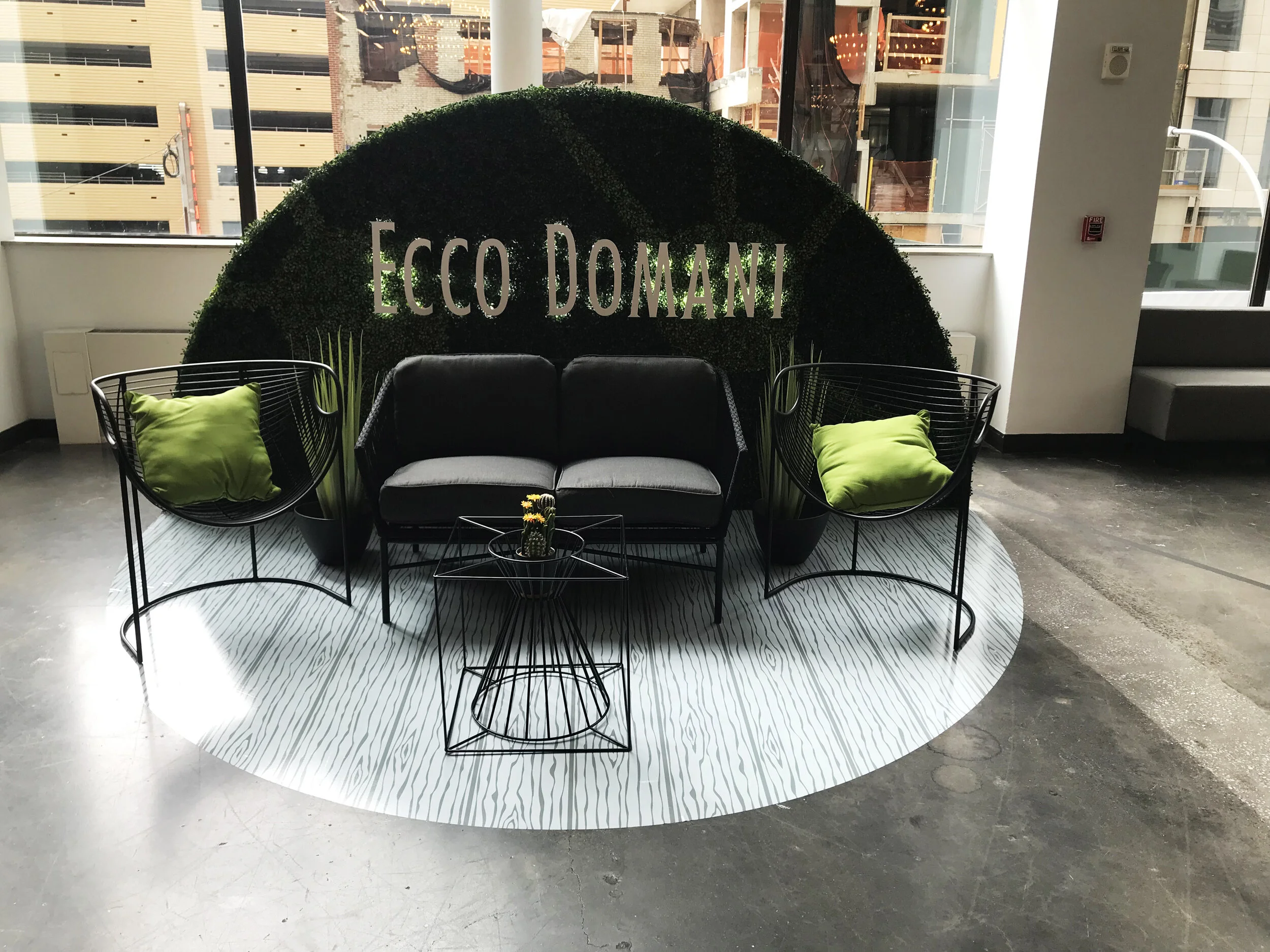 Modern indoor lounge area with black sofa, two black wire chairs with lime green pillows, round black wire side table with a small cactus, and a patterned white and black rug, with a green wall sign that says "Ecco Duman" behind the seating.