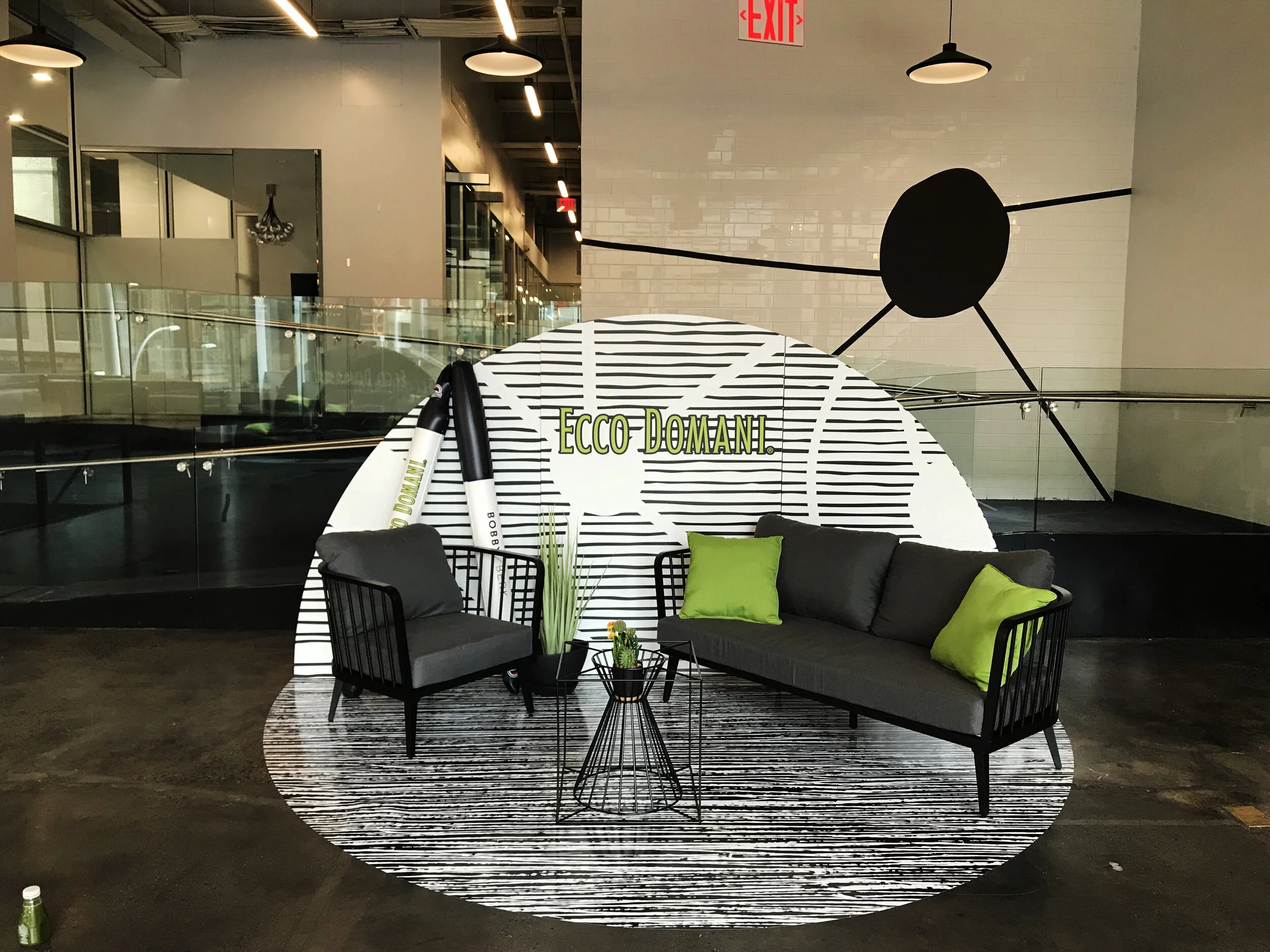 Indoor seating area with black chairs, a gray couch with green pillows, and a black wire side table with plants. A black-and-white striped rug is on the floor, and a white circular backdrop with black lines and the text "ECCO DOMANI" in green is in t
