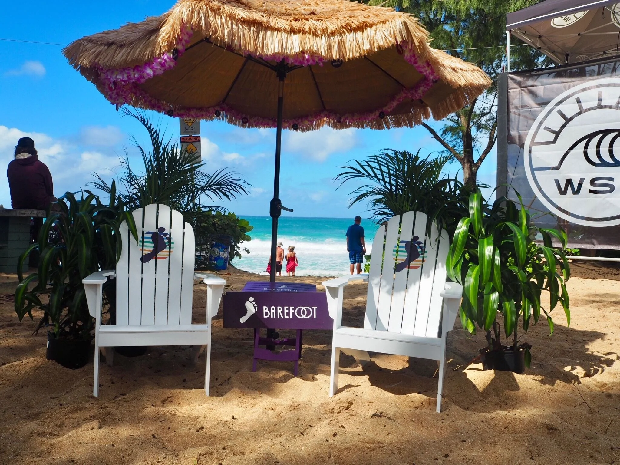 A daytime beach scene viewed through a thatched umbrella with pink flower decorations. There are two white Adirondack chairs in the foreground with a barefoot logo and the word "Barefoot" on a sign. In the background, people are walking along the san