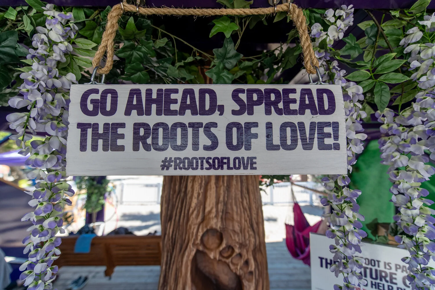 A wooden sign with purple lettering that reads, 'Go ahead, spread the roots of love! #RootsofLove', hanging from a rope among lush green leaves and purple flowers.