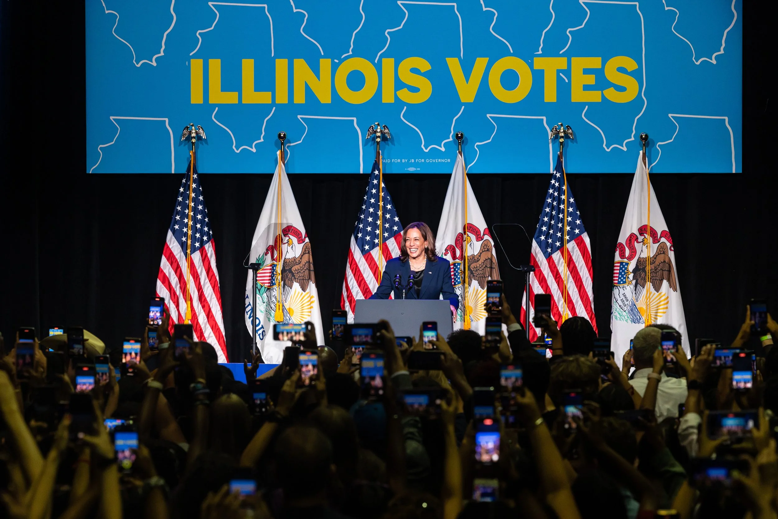 Vice President Kamala Harris speaking at the Illinois Gubernatorial Election Night event with flags of Illinois and the United States behind her and a large banner that reads 'Illinois Votes' in the background, with a crowd taking pictures.