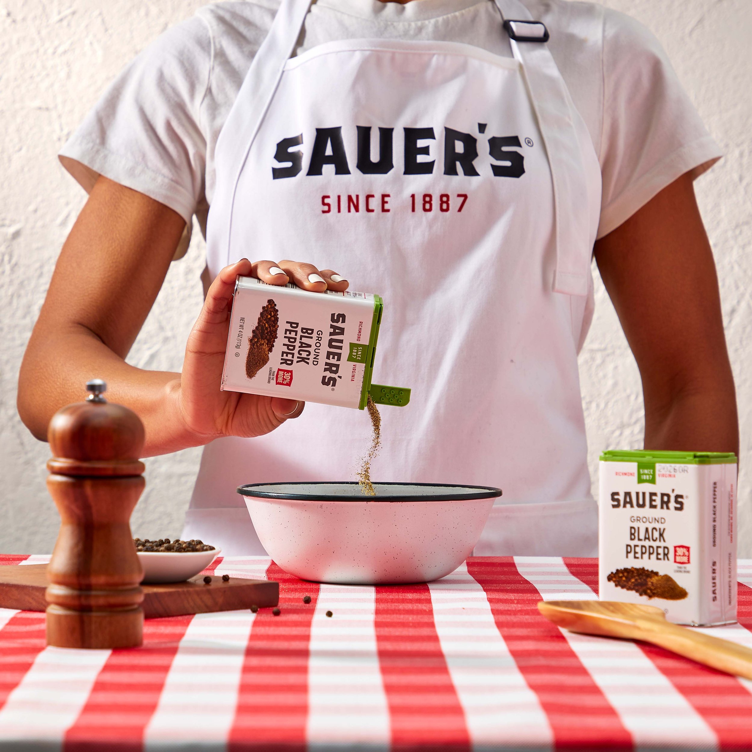 Person standing at a table with a red and white checkered tablecloth, holding a box of Sauer's ground black pepper, with a white apron and a gray shirt, in front of a white textured wall.