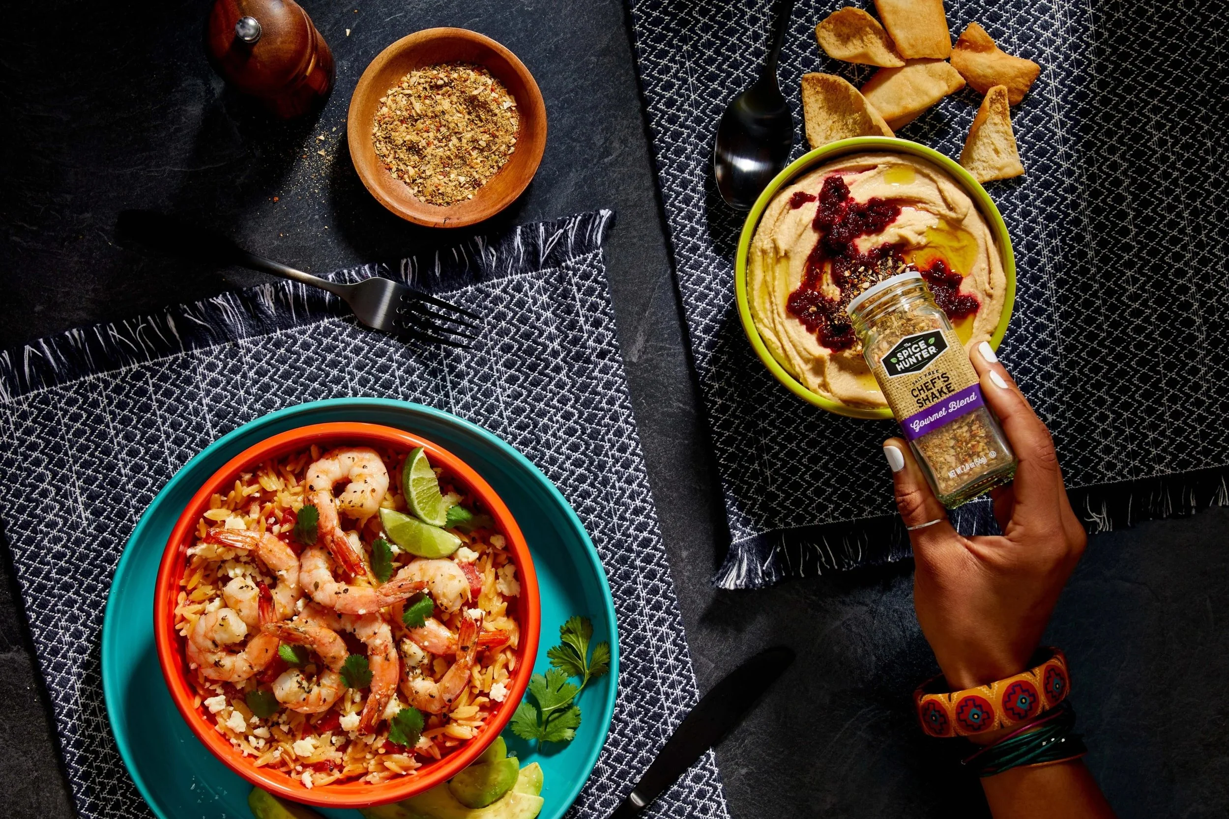 A vibrant blue bowl filled with shrimp and rice, garnished with cilantro and lime wedges, on a black table with a black and white patterned cloth. Part of a dish with sliced bread, and a round yellow bowl with hummus topped with red berry sauce and o