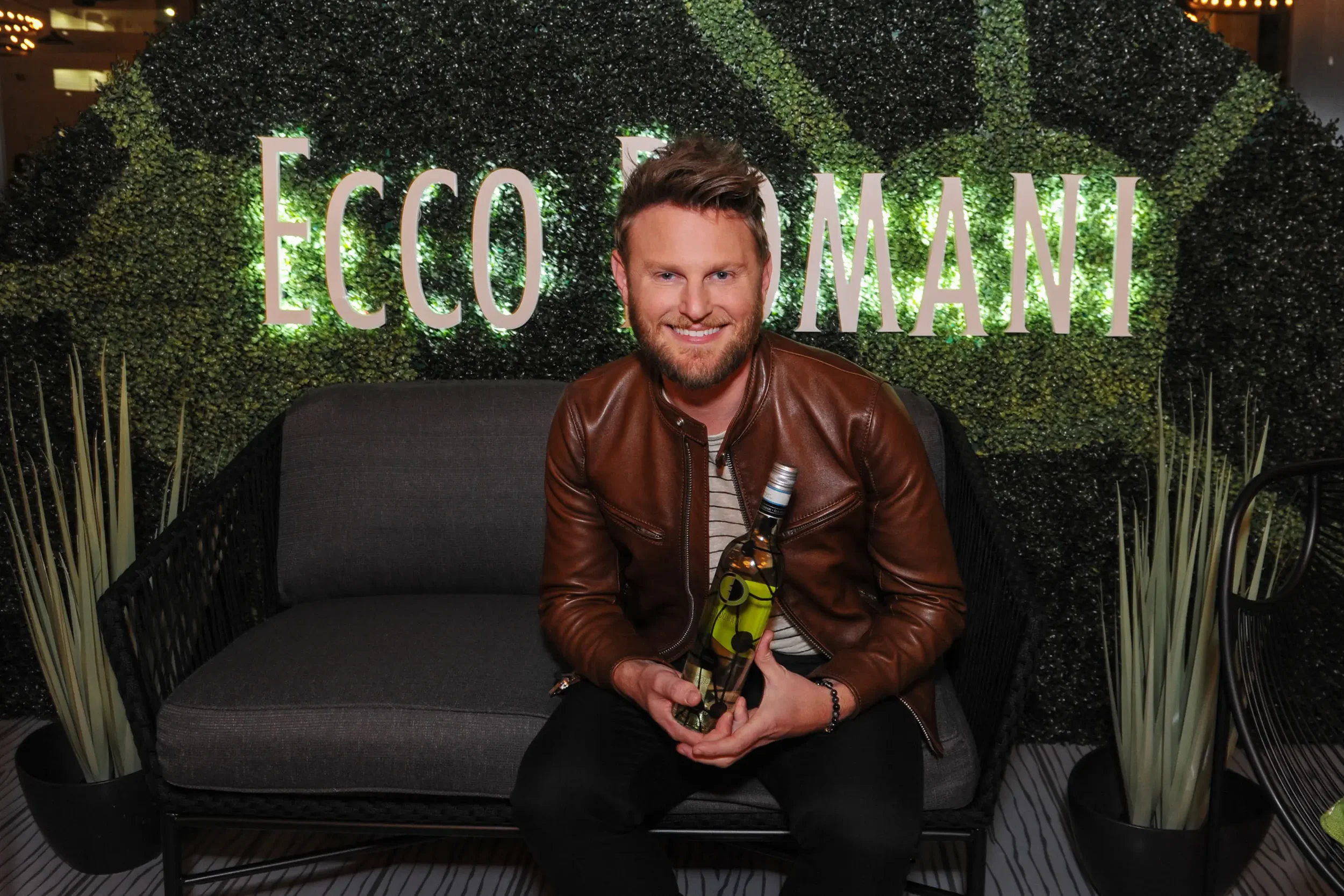 A man smiling and sitting on a black outdoor sofa, holding a bottle of beer and sunglasses, in front of a hedge wall with the word 'ECO' and part of another word illuminated behind him.