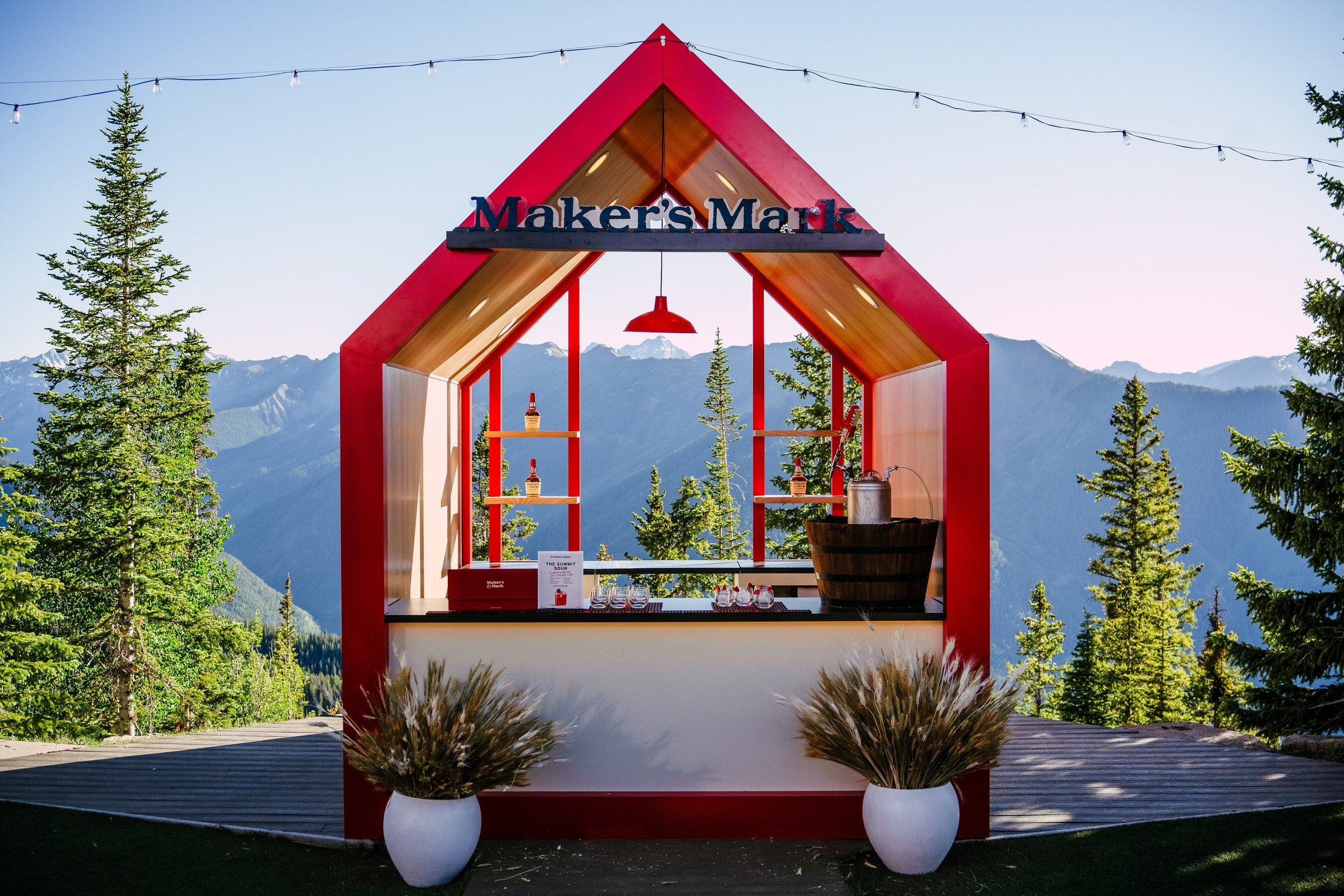 Outdoor bar setup at Maker's Mark with red and wooden arch, mountains in background, string lights, and potted plants.