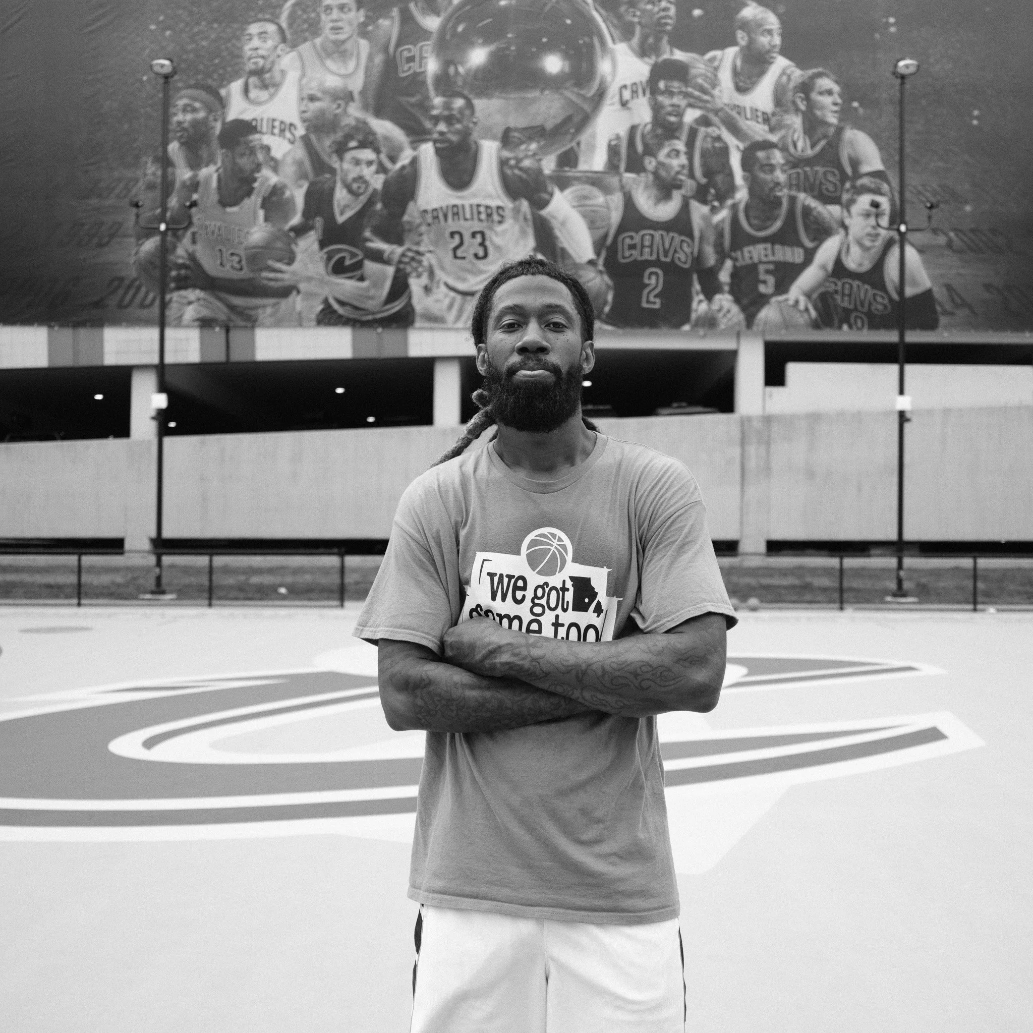 A man with a beard, wearing a T-Shirt with a basketball logo, stands in front of a large black-and-white poster of a basketball team, with arms crossed.