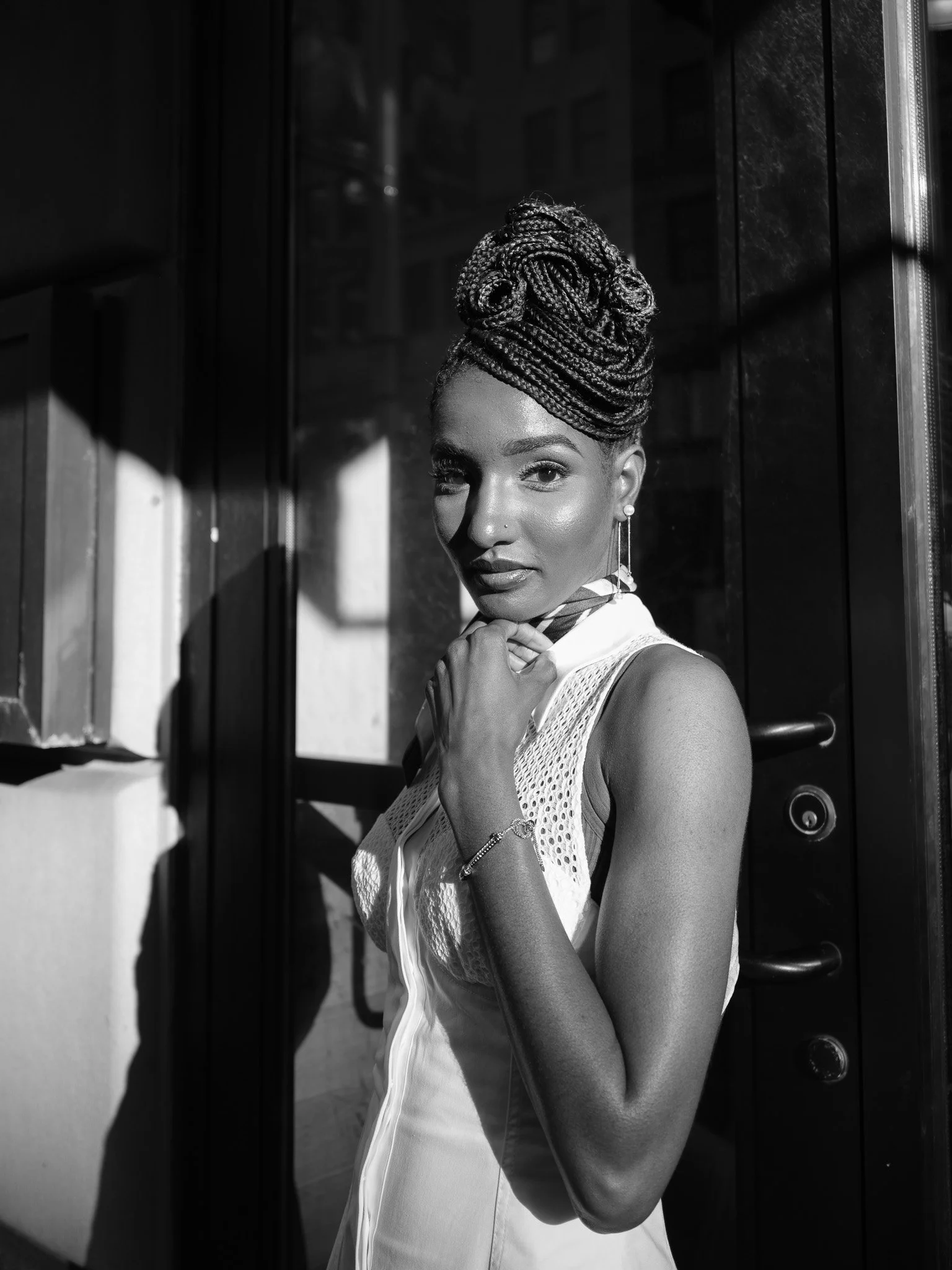 Black and white photo of a black woman with braided hair, softly looking at the camera, standing outside near a building with shadows. She wears earrings, a watch, and a sleeveless top.