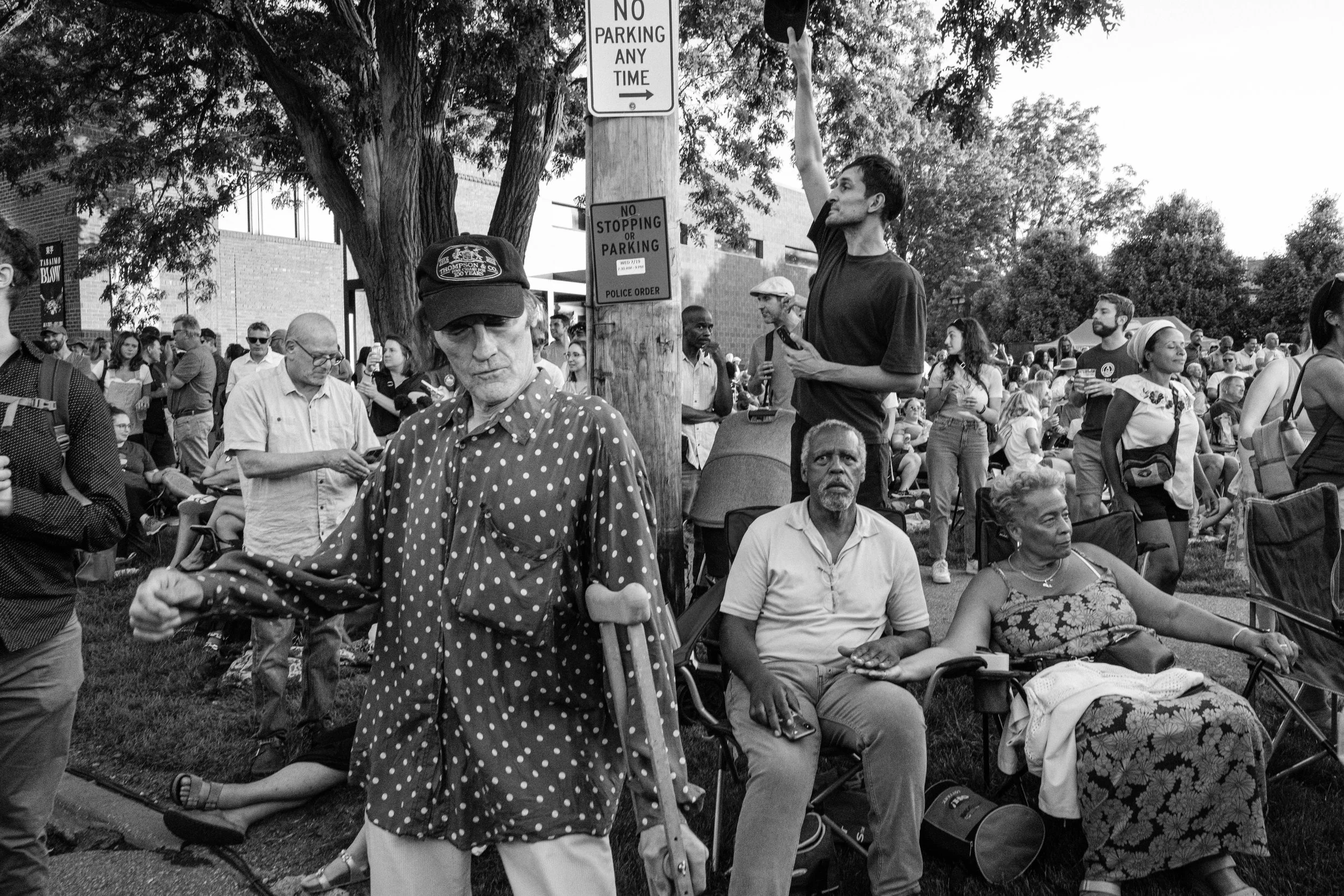 A crowd of people sitting and standing outdoors, with some chairs and trees, during an event or gathering in black and white.