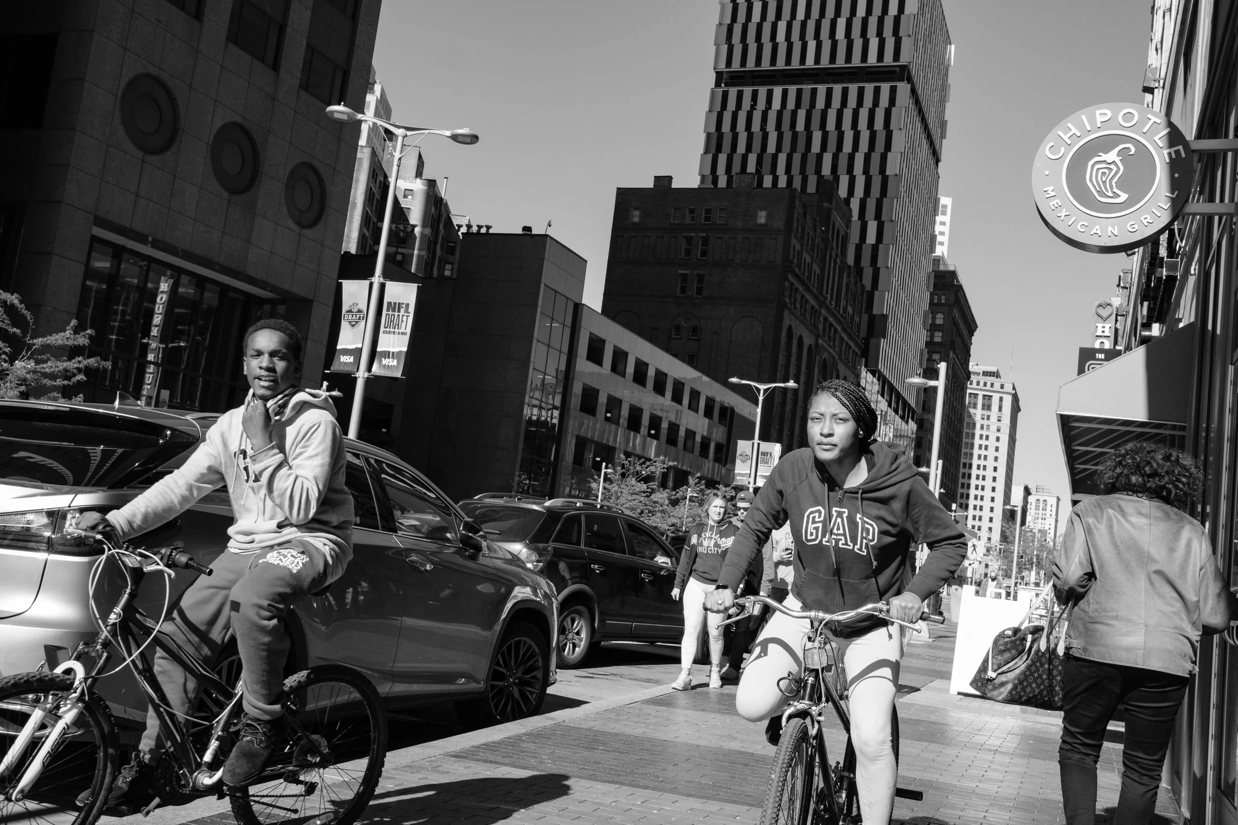 People riding bicycles and walking on a city street with tall buildings and a Chipotle Mexican Grill sign in the background on a sunny day.