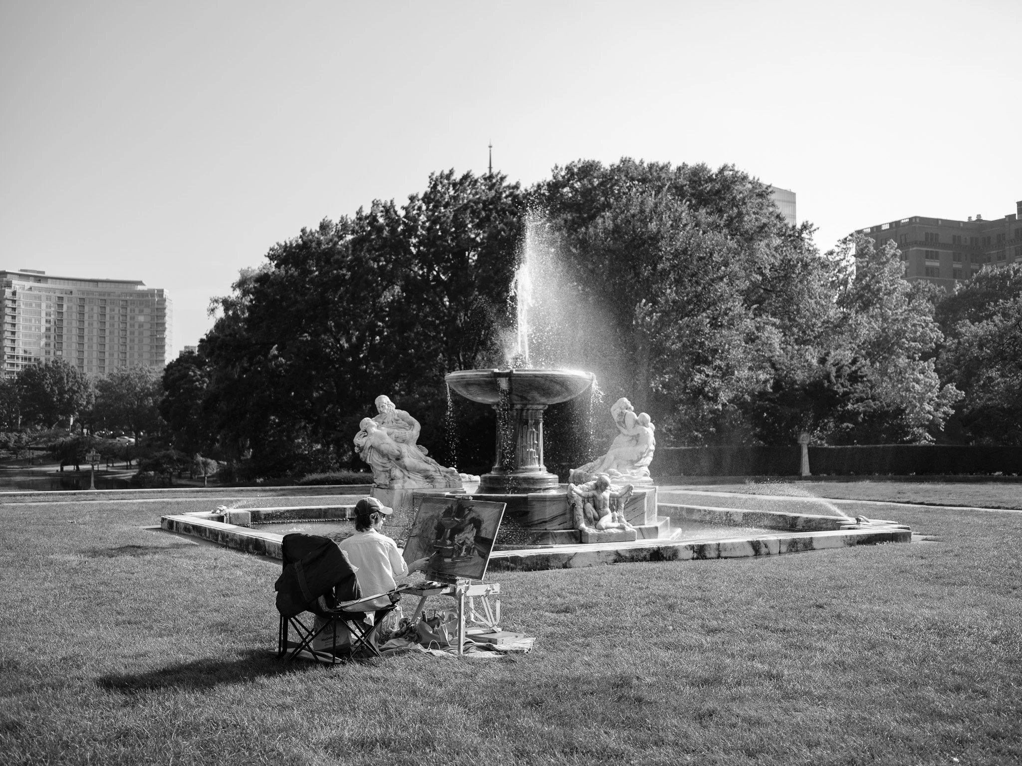 A man painting a fountain sculpture in a park with trees, modern buildings, and a clear sky in the background.