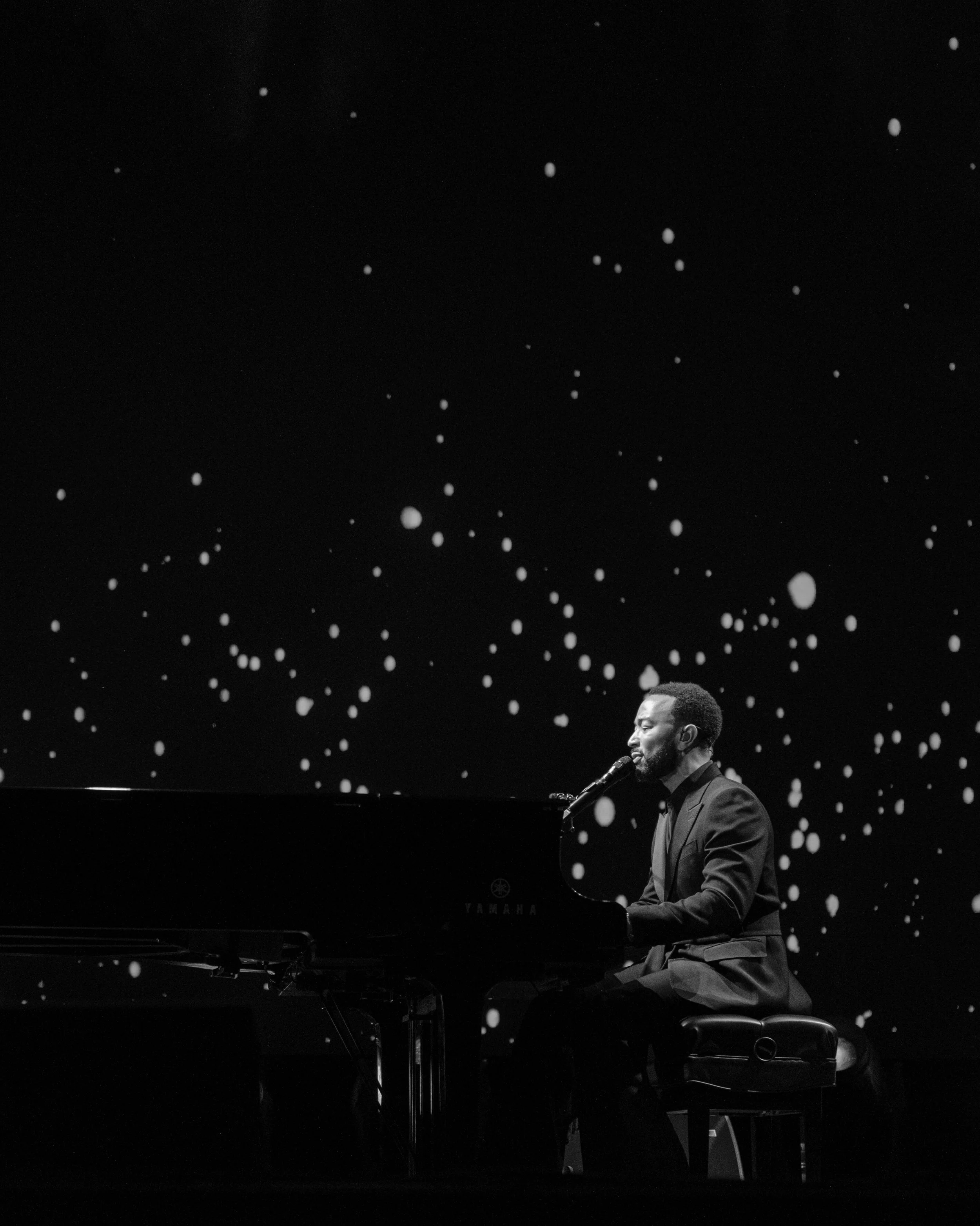 John Legend playing his piano on a dimly lit stage with a starry background.