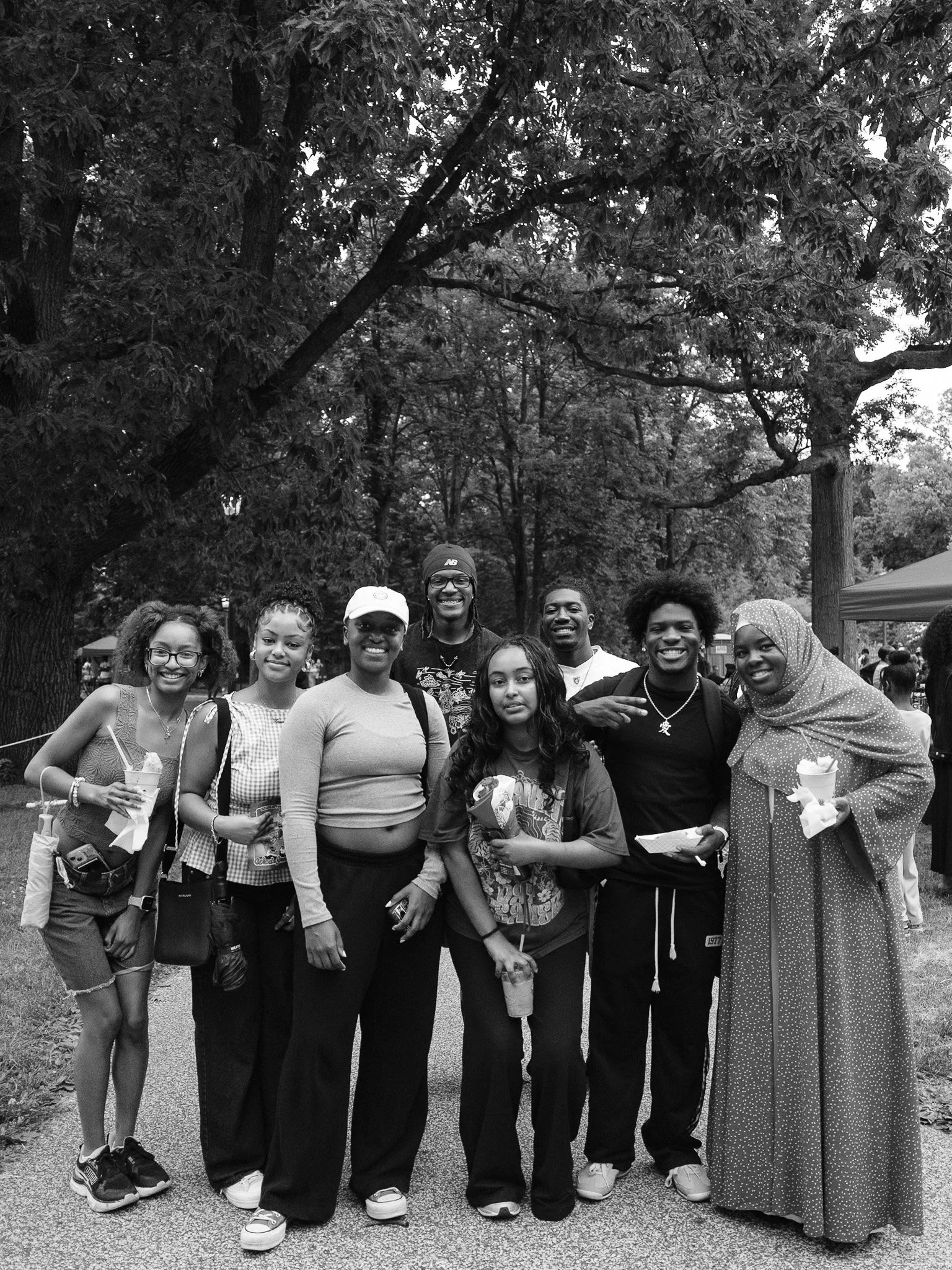 A group of diverse people smiling at an outdoor event, holding drinks, with trees and tents in the background.