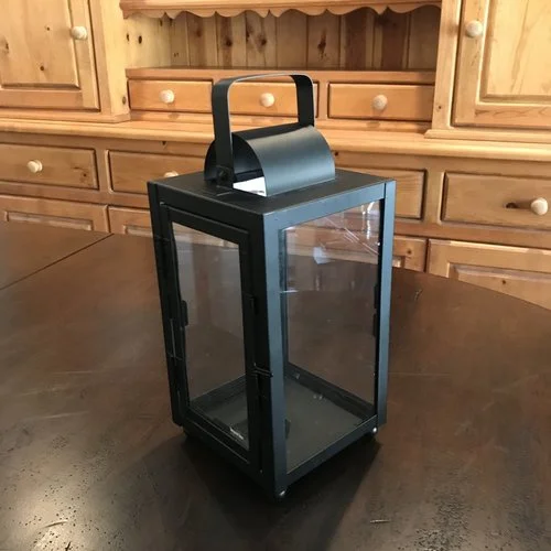 Small Black Lantern Matte black metal lantern with glass sides. Openable door on one side. Handle can be used for hanging. 12" tall x 6" square. Comes with three candles in glass.