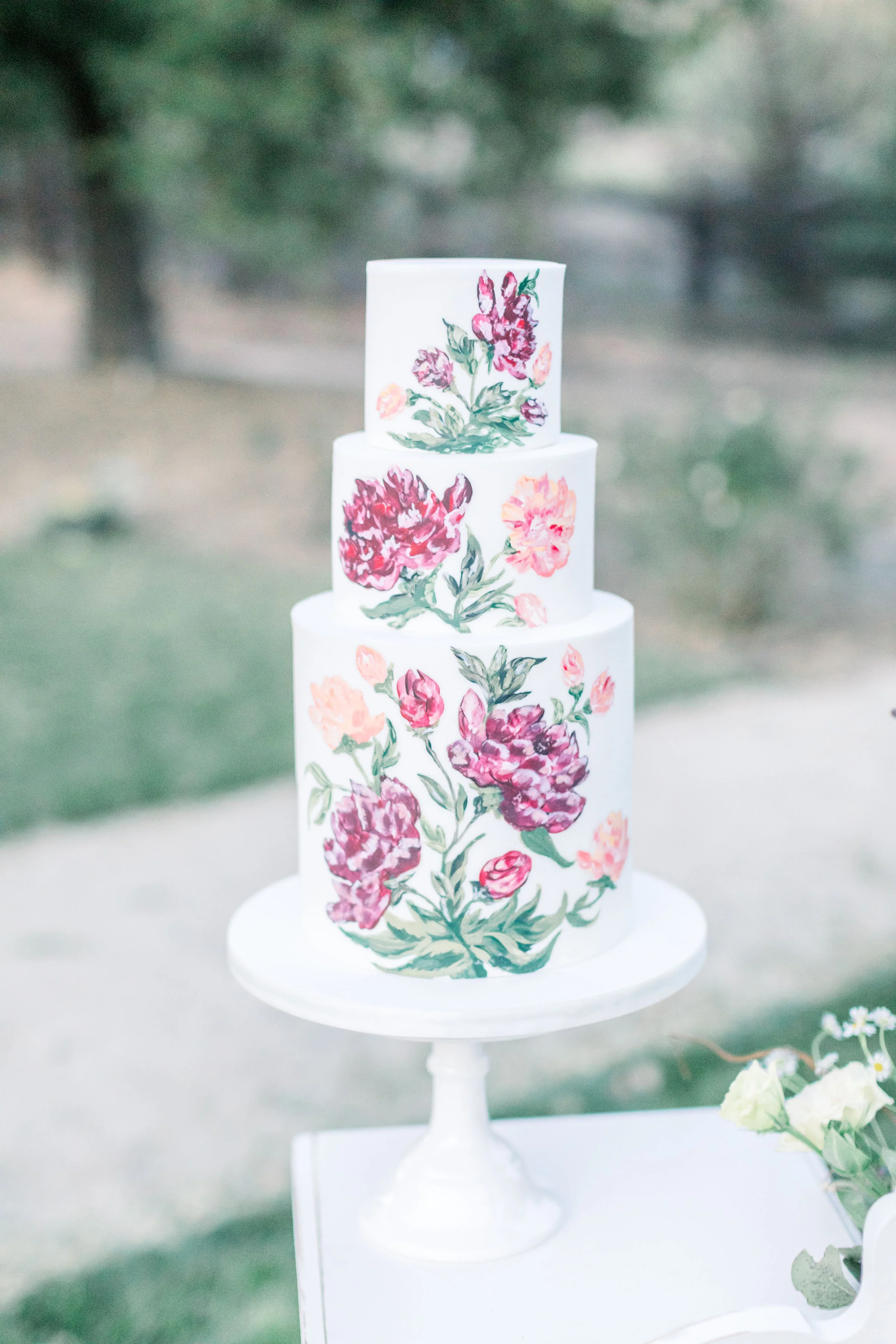 A hand painted floral wedding cake in pink tones. Three tiered white base frosting. White pedestal.