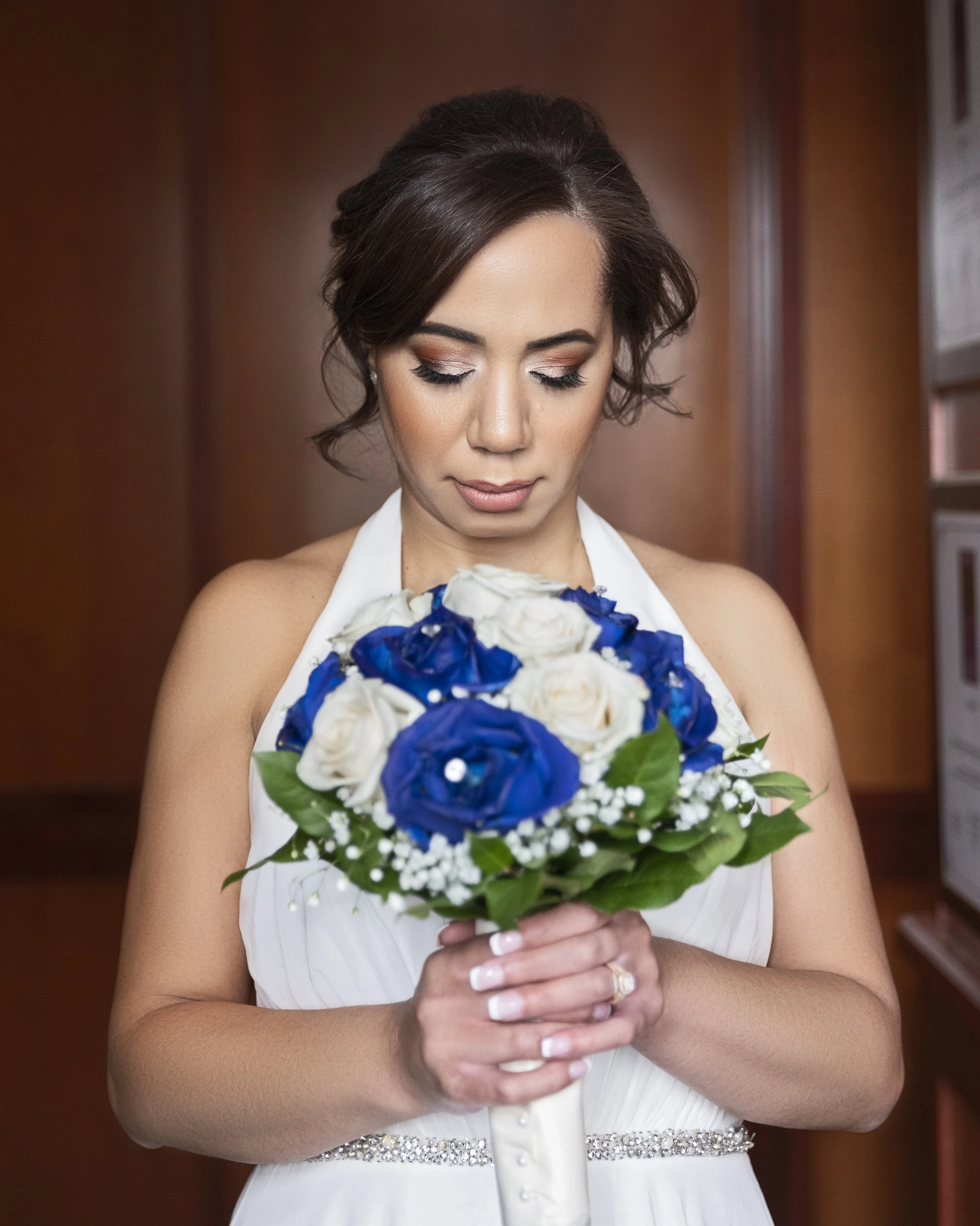 Bride holding a bouquet of blue and white roses, wearing a white dress with a halter neck and rhinestone belt, eyes closed.