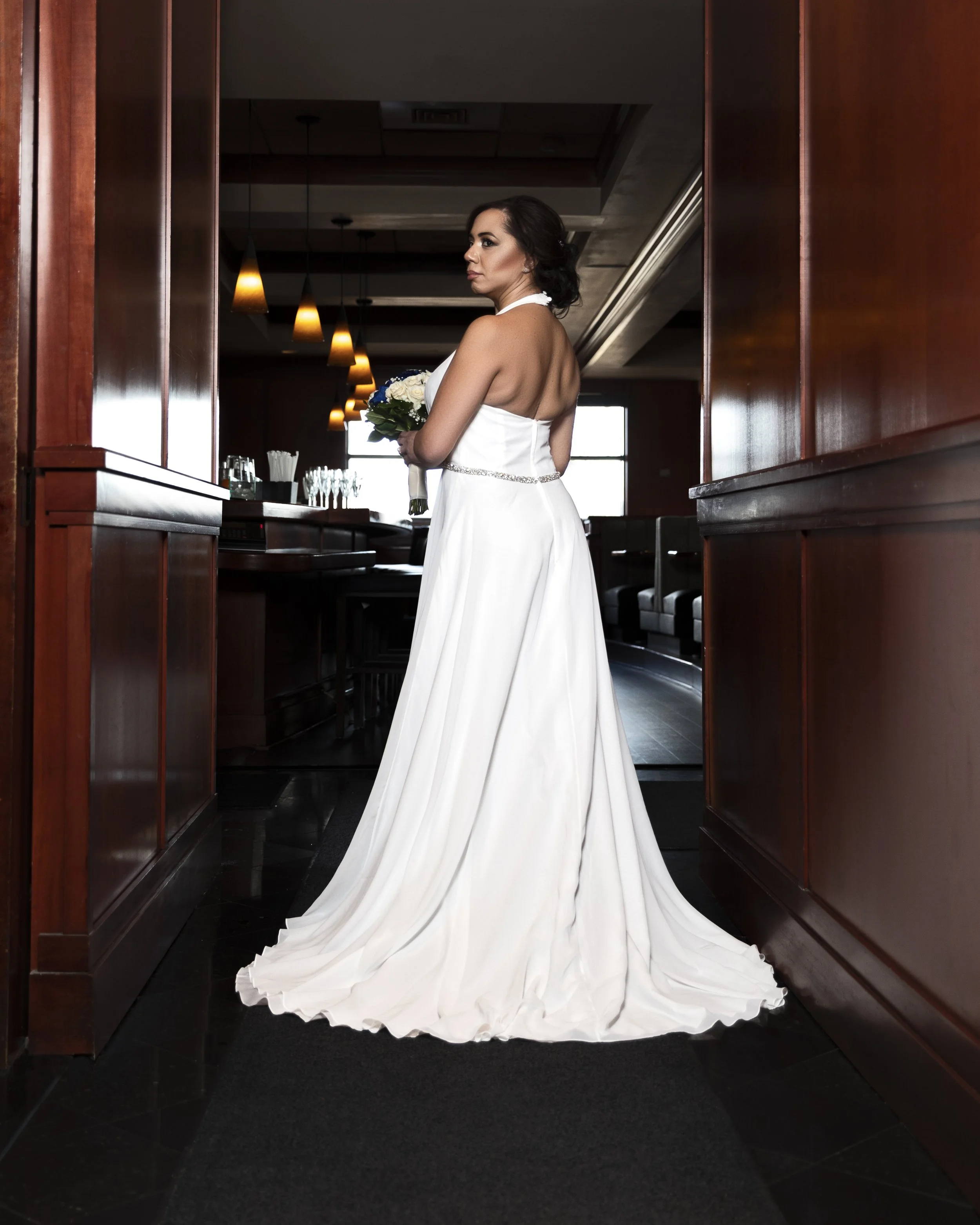 Bride in a white wedding dress holding a bouquet, standing in a dimly-lit hallway with wooden paneling and pendant lights.