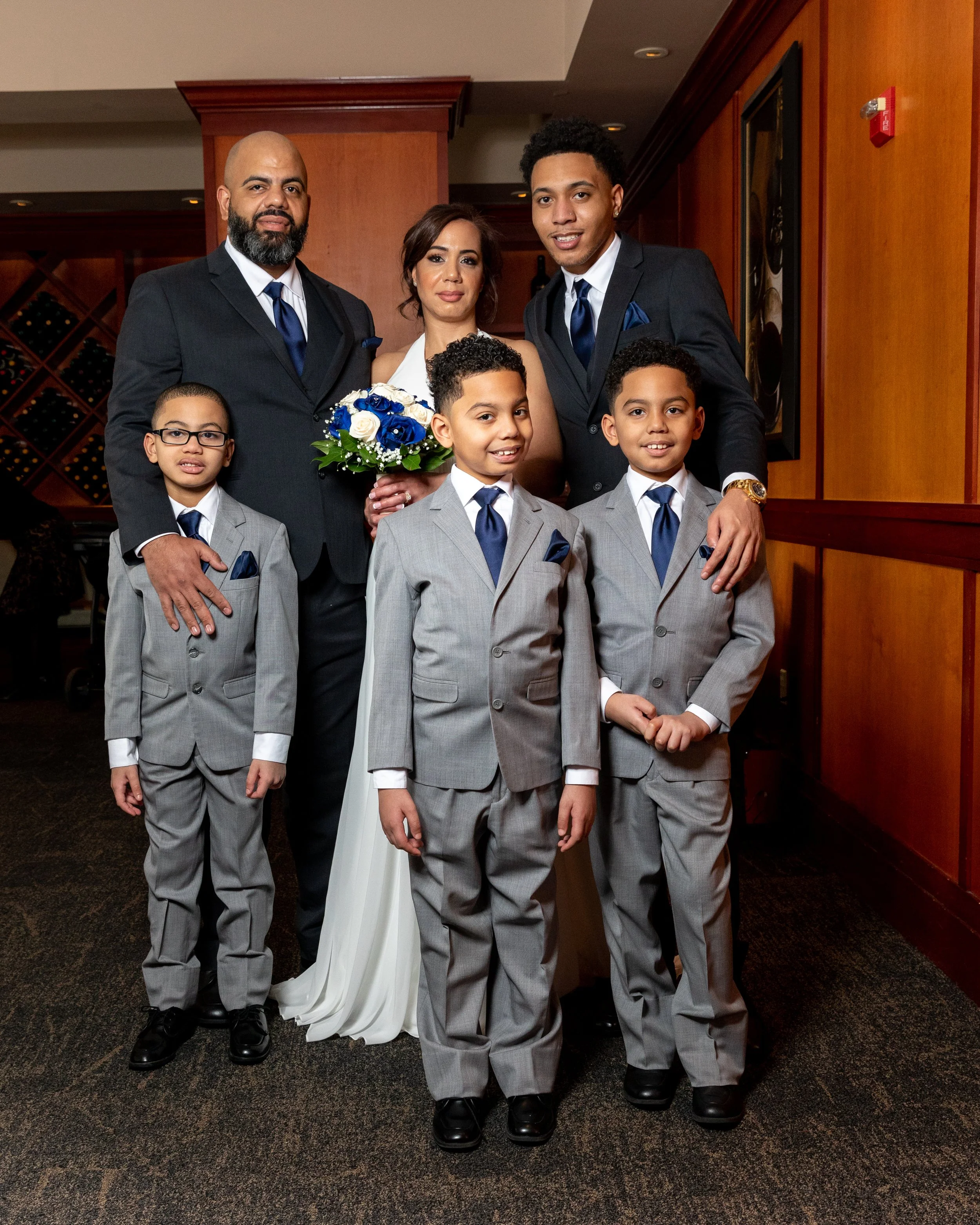 A family photo with a couple in formal wear standing with four children also dressed in suits. The woman is holding a bridal bouquet.