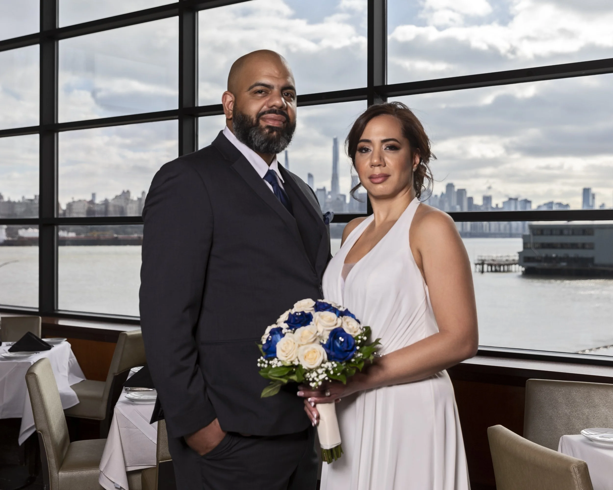 A couple standing indoors by a large window, with a city skyline in the background. The woman is in a white dress holding a bouquet with blue and white flowers. The man is wearing a dark suit. The area has a dining setup with tables and chairs.