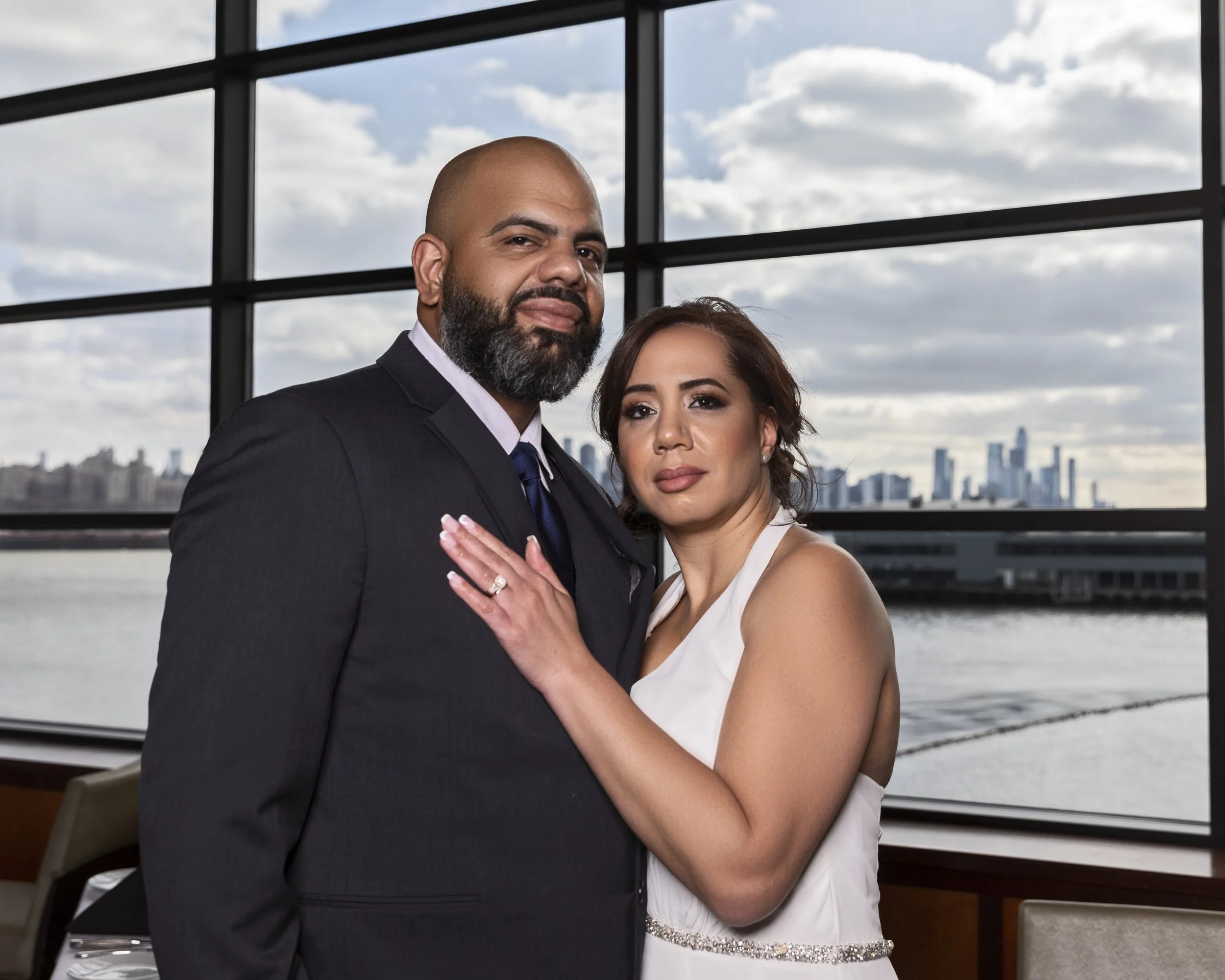 A couple posing together indoors, with a city skyline and water visible through large windows in the background. The woman is wearing a white dress and the man is in a suit.