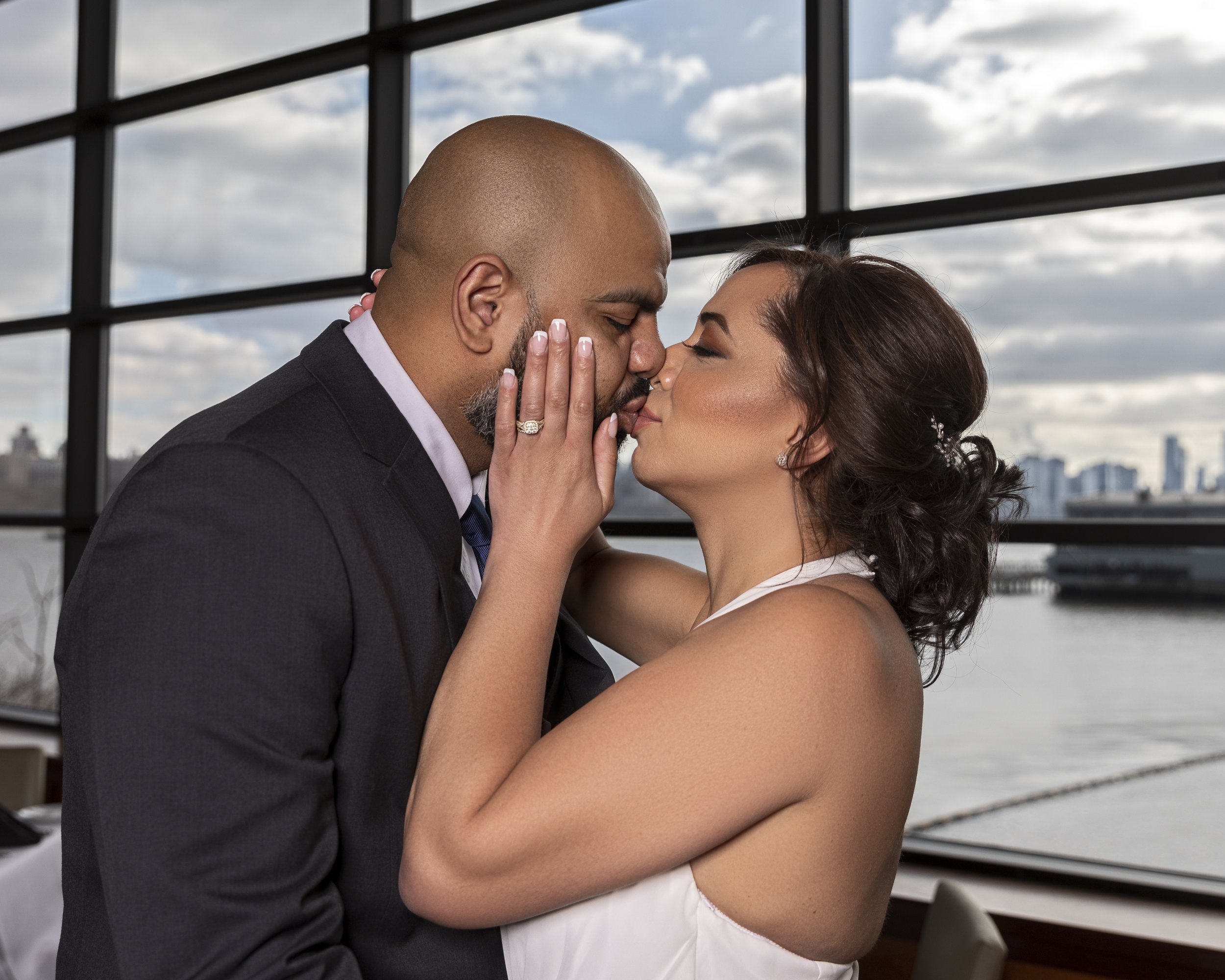 A couple sharing a kiss, with the woman wearing a white dress and an engagement ring. They are standing indoors near large windows overlooking a cityscape and water.