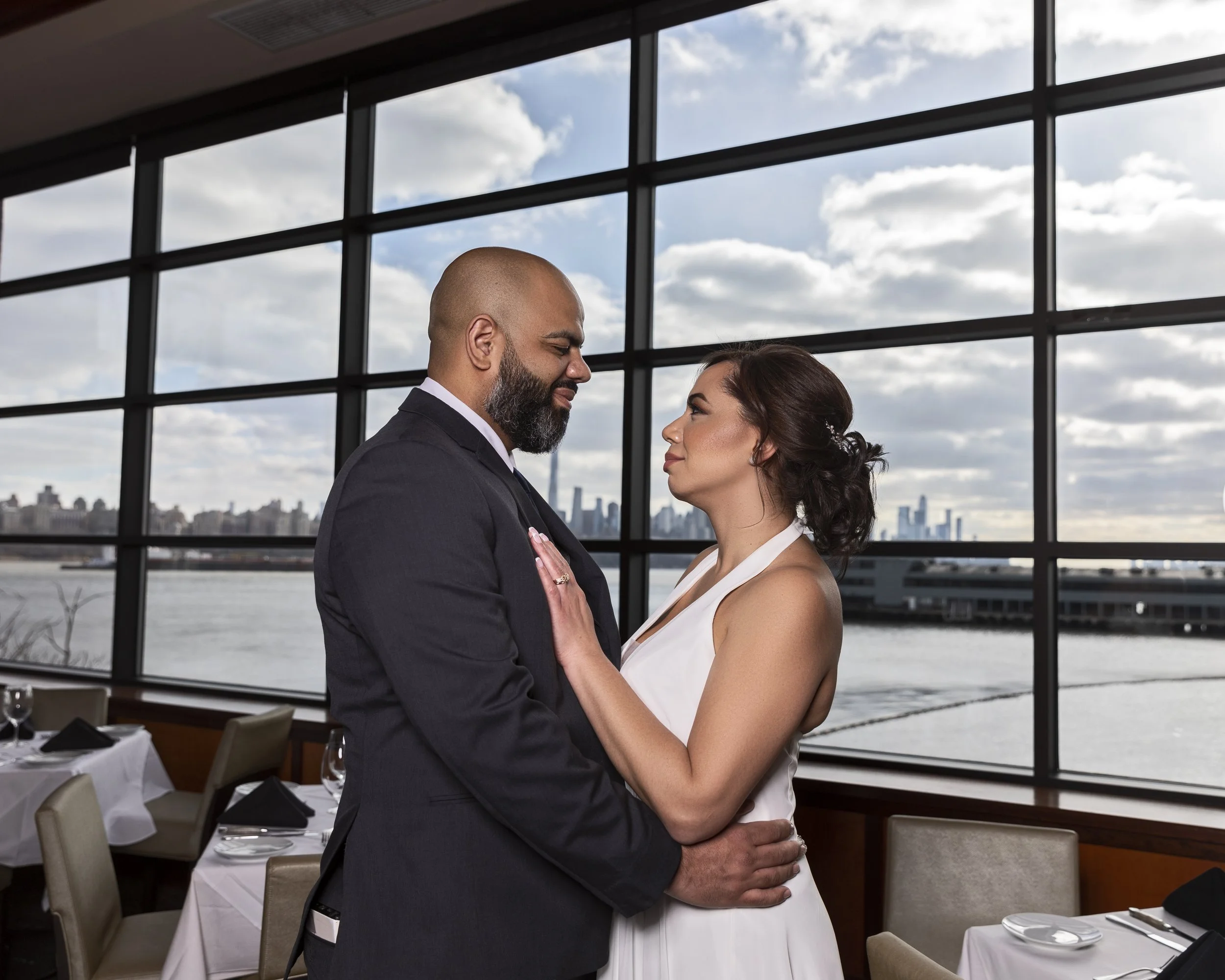 A couple in formal attire embracing in a restaurant with a large window overlooking a water view and cityscape.