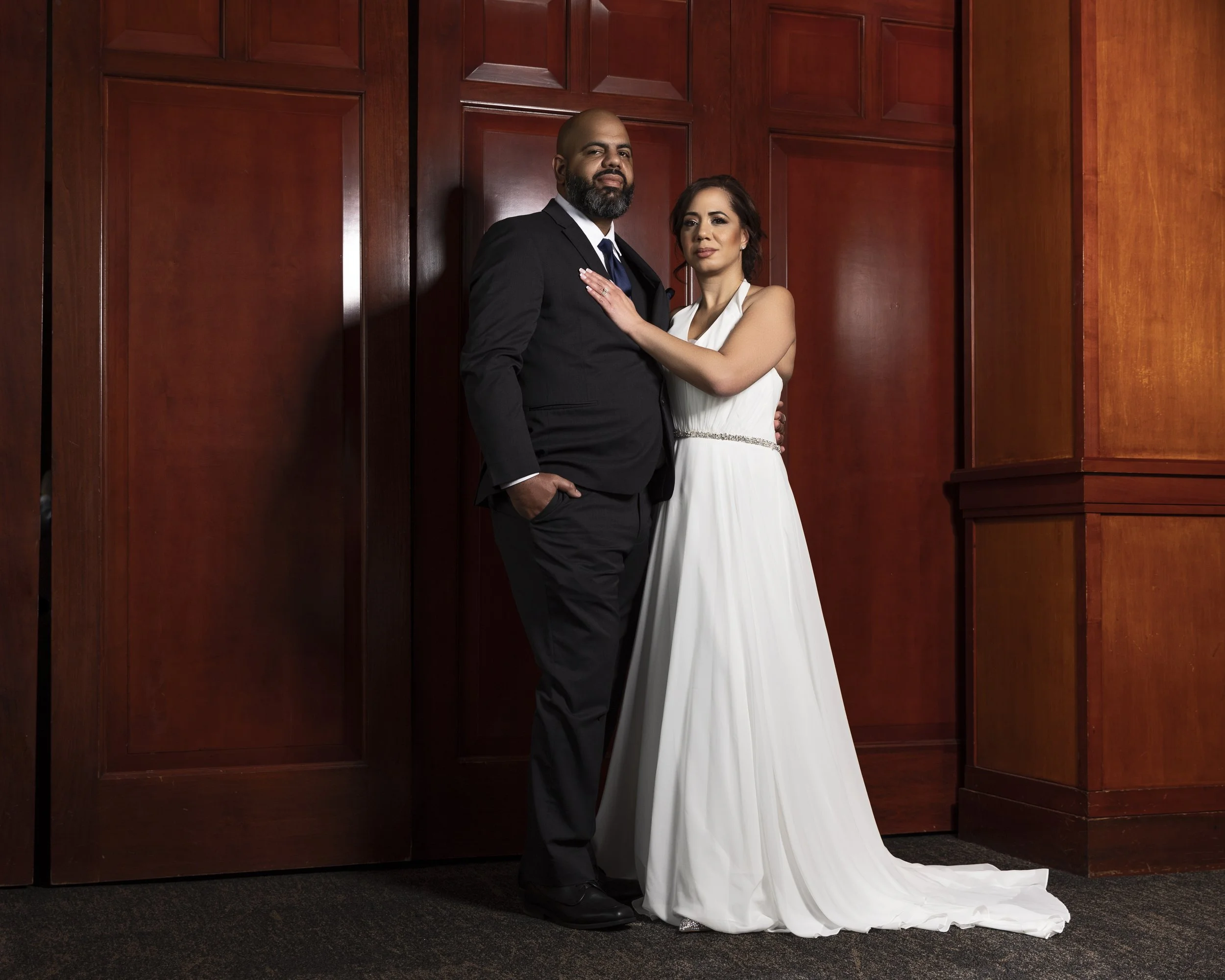 A couple posing together, with the man wearing a black suit and the woman in a white gown, standing in front of wooden paneling.