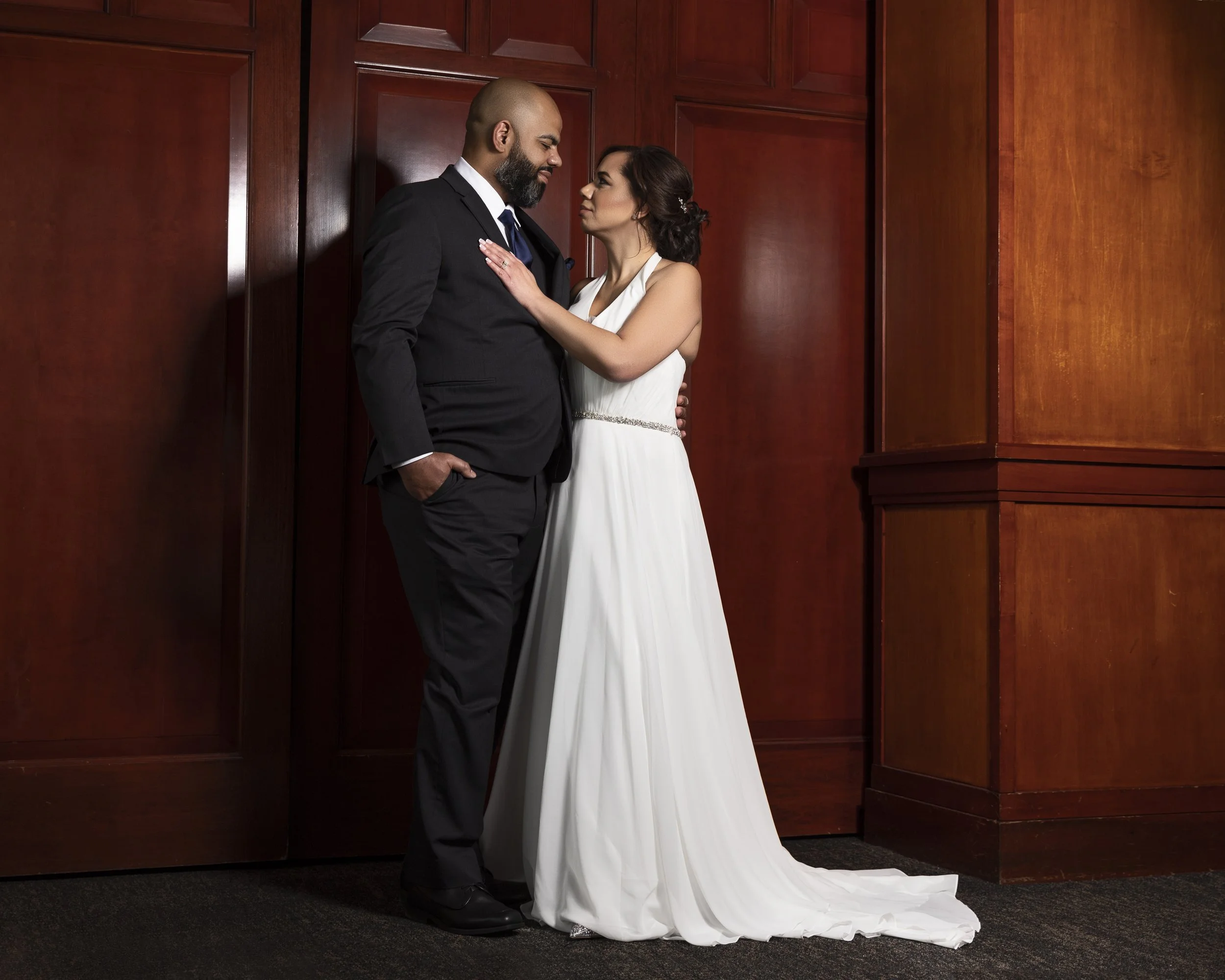 Bride in white gown and groom in dark suit standing close in front of a wooden wall.