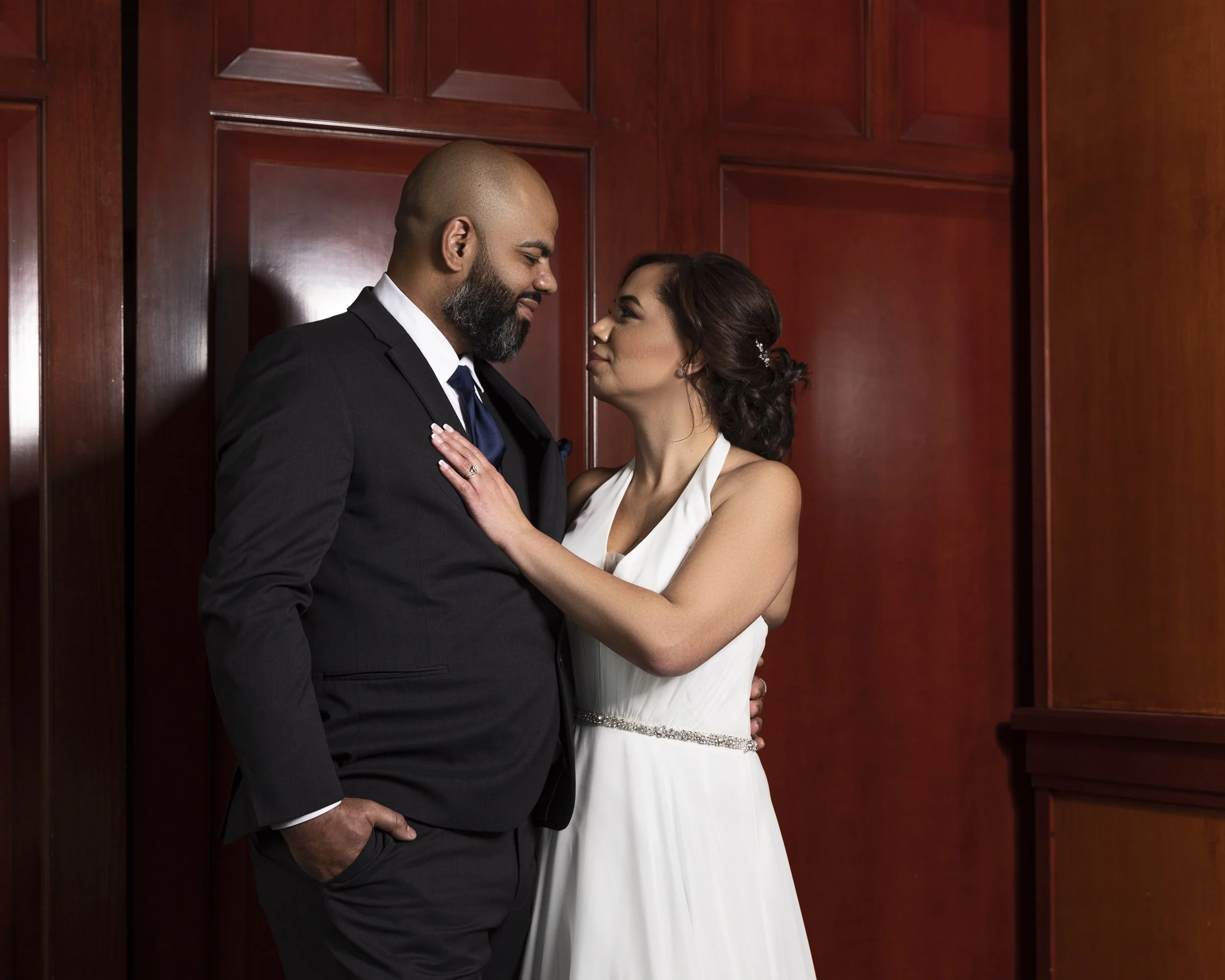 Bride in white dress and groom in black suit, embracing in front of a wooden paneled wall.