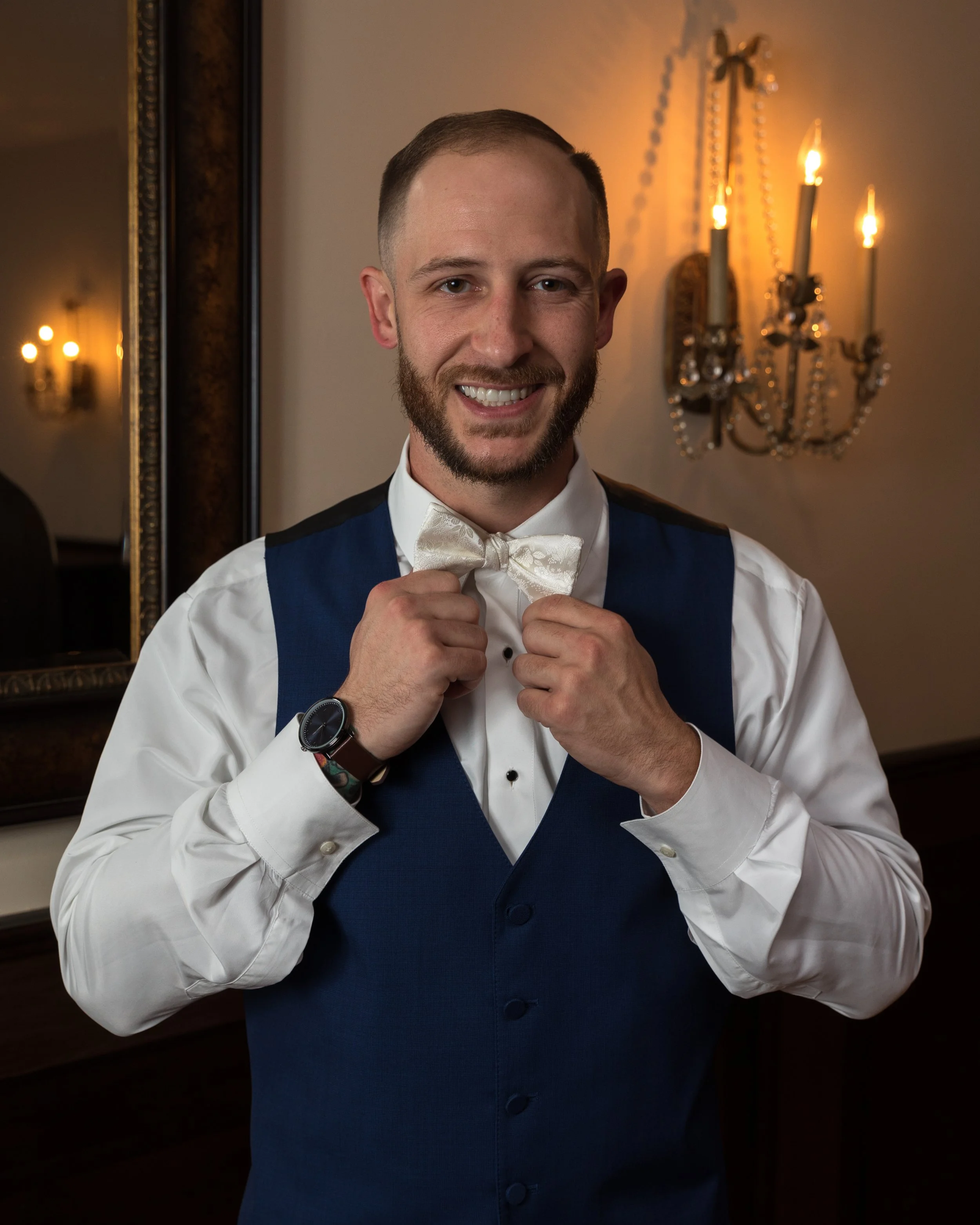 Man in formal attire, adjusting bow tie, smiling, standing in front of a mirror with chandelier lighting in the background.