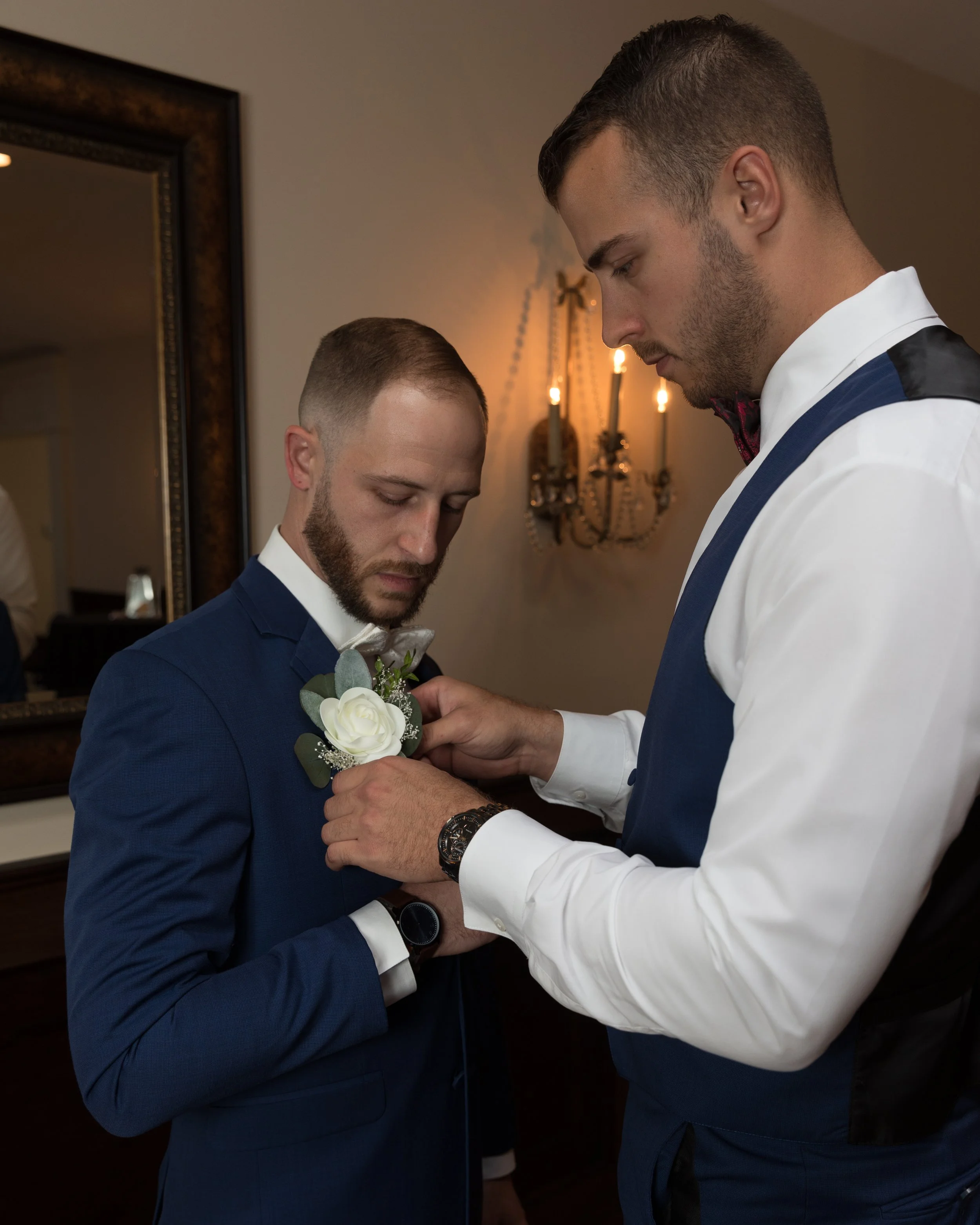 Two men in suits, one helping the other with a boutonniere, indoor setting with mirror and wall sconce.