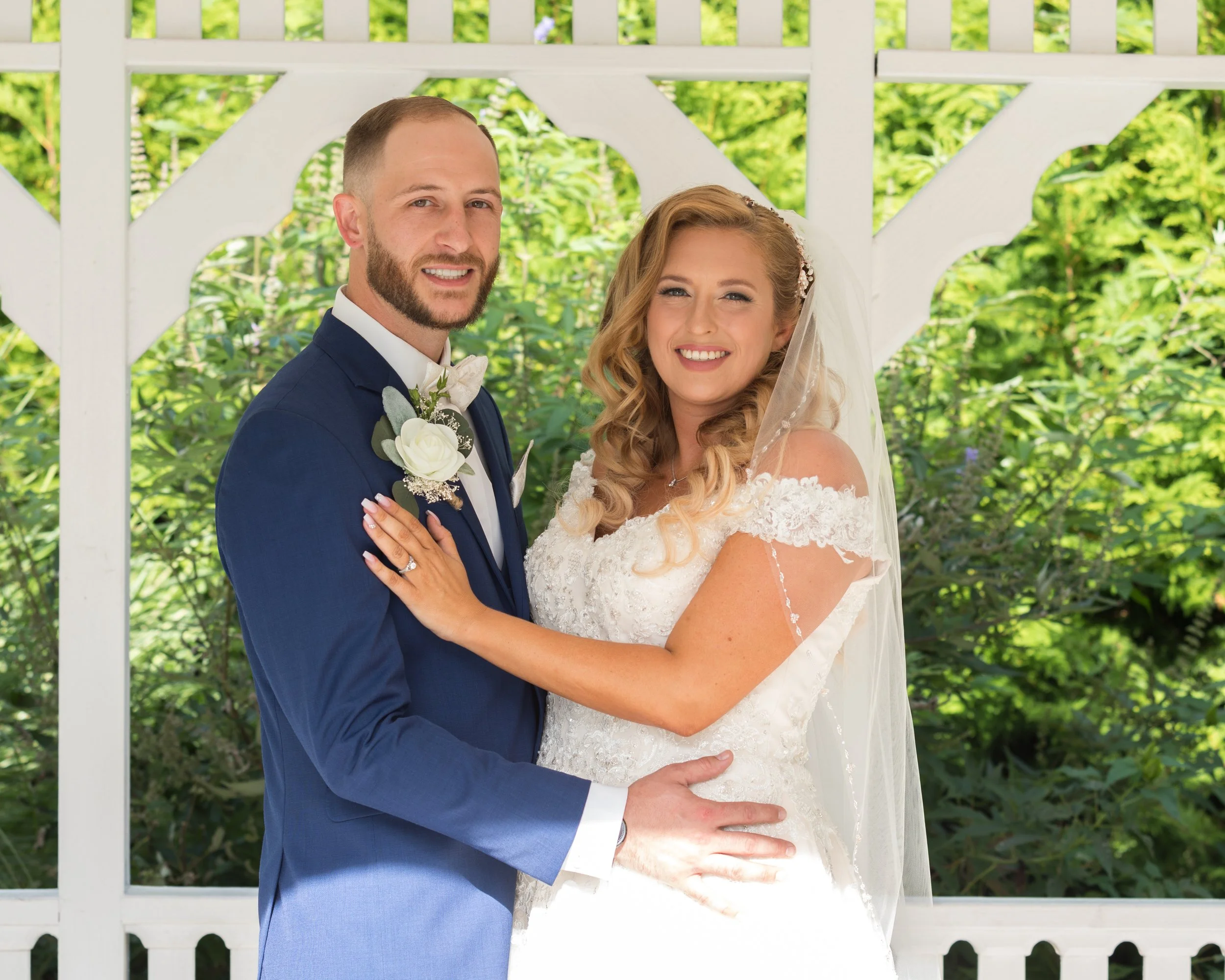 Bride and groom posing in a garden gazebo, with bride wearing a white gown and veil, and groom in a blue suit and white boutonniere.