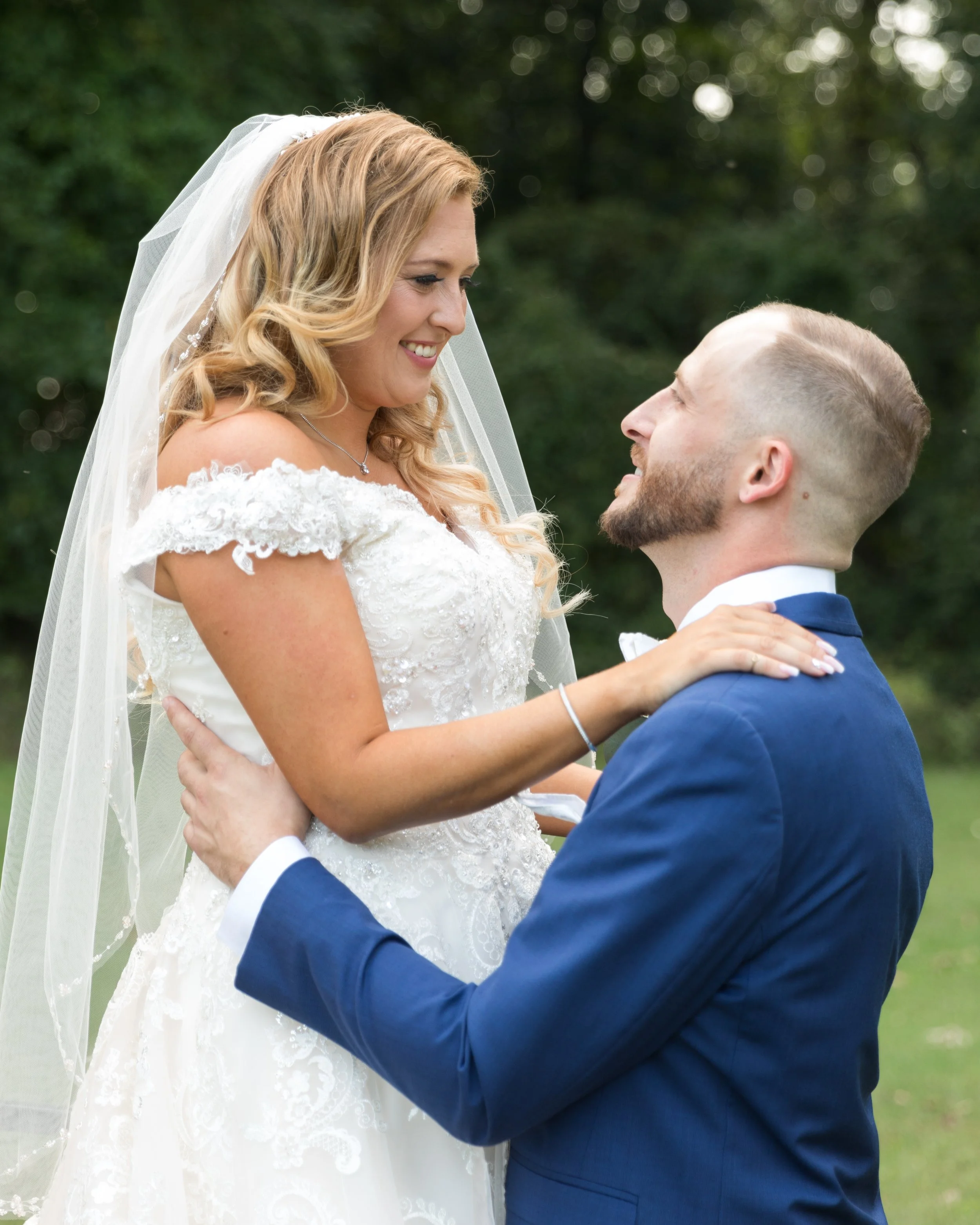A bride and groom smiling at each other. The bride is wearing a white wedding dress and veil, while the groom is in a blue suit. They are outdoors with greenery in the background.