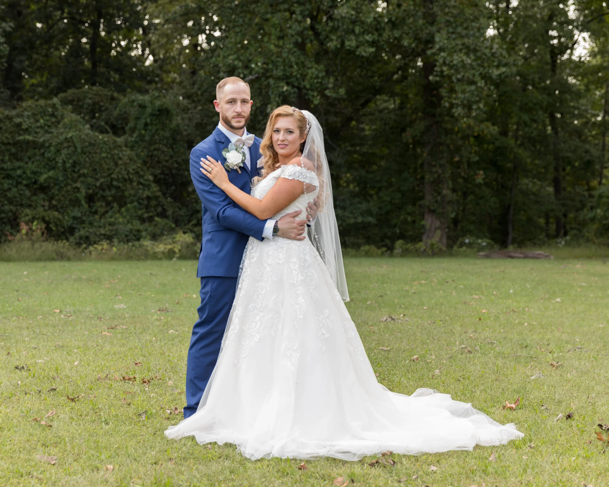Bride and groom in wedding attire standing in a grassy field with trees in the background.