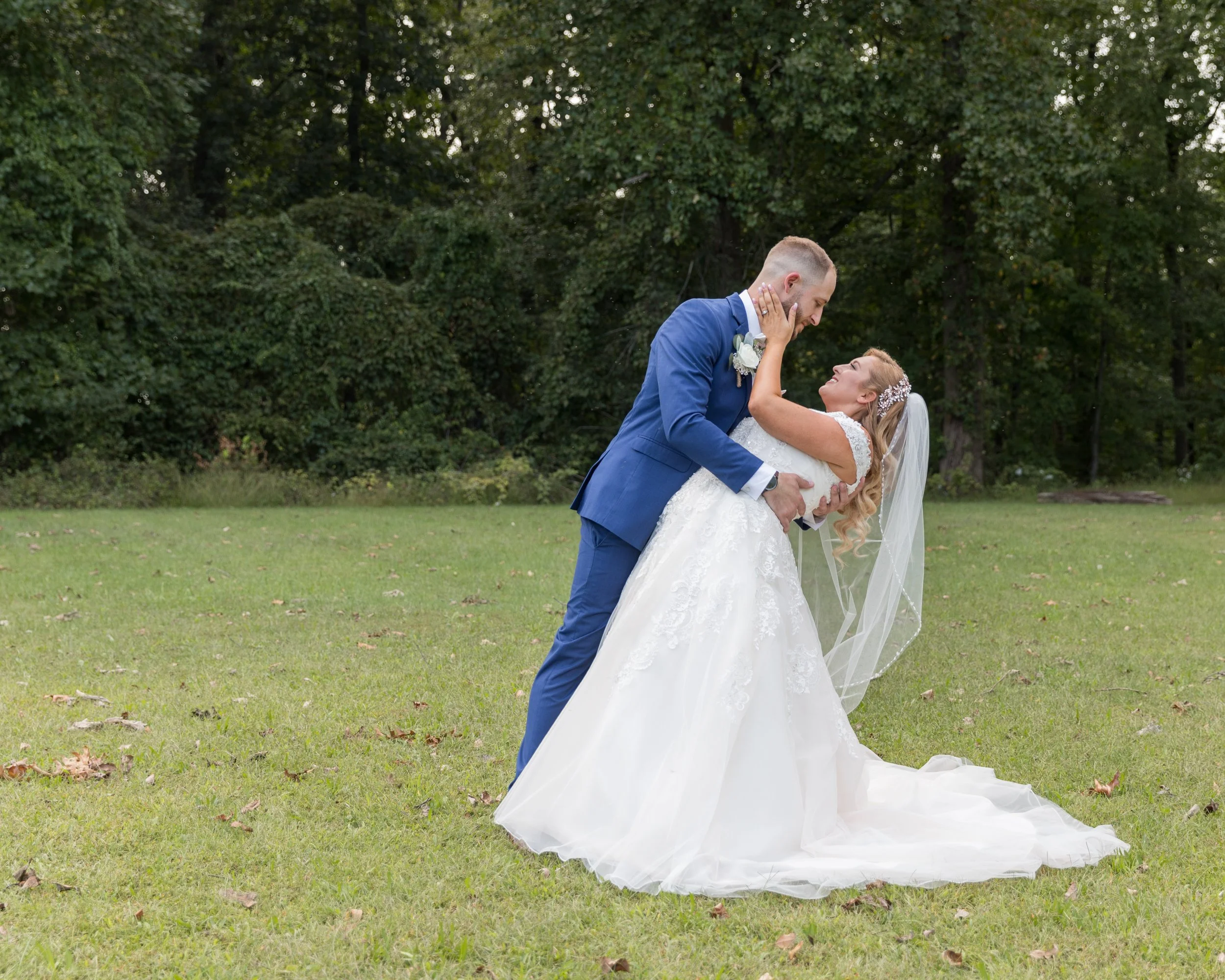 Bride and groom dancing outdoors in a grassy area, surrounded by trees, with the groom in a blue suit and the bride in a white gown and veil.