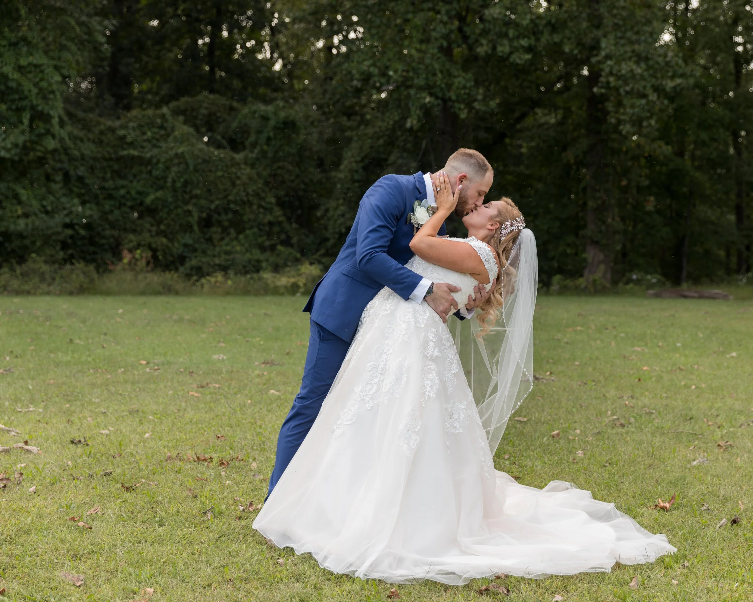 A bride in a white wedding dress and veil being kissed by a groom in a blue suit, outdoors in a grassy area with trees in the background.