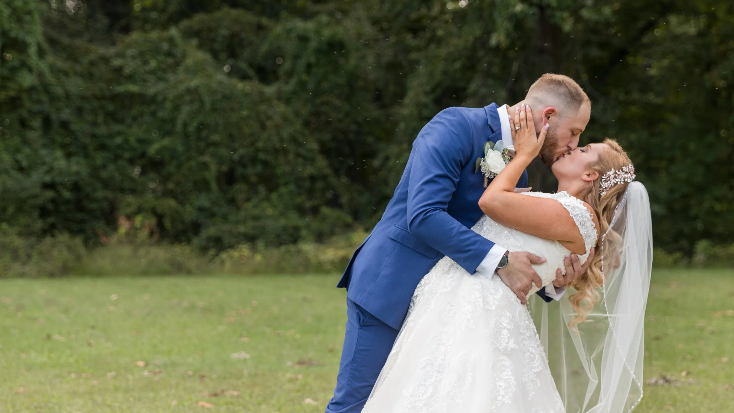 Bride and groom kissing outdoors, groom in blue suit, bride in white wedding dress with veil, green background.
