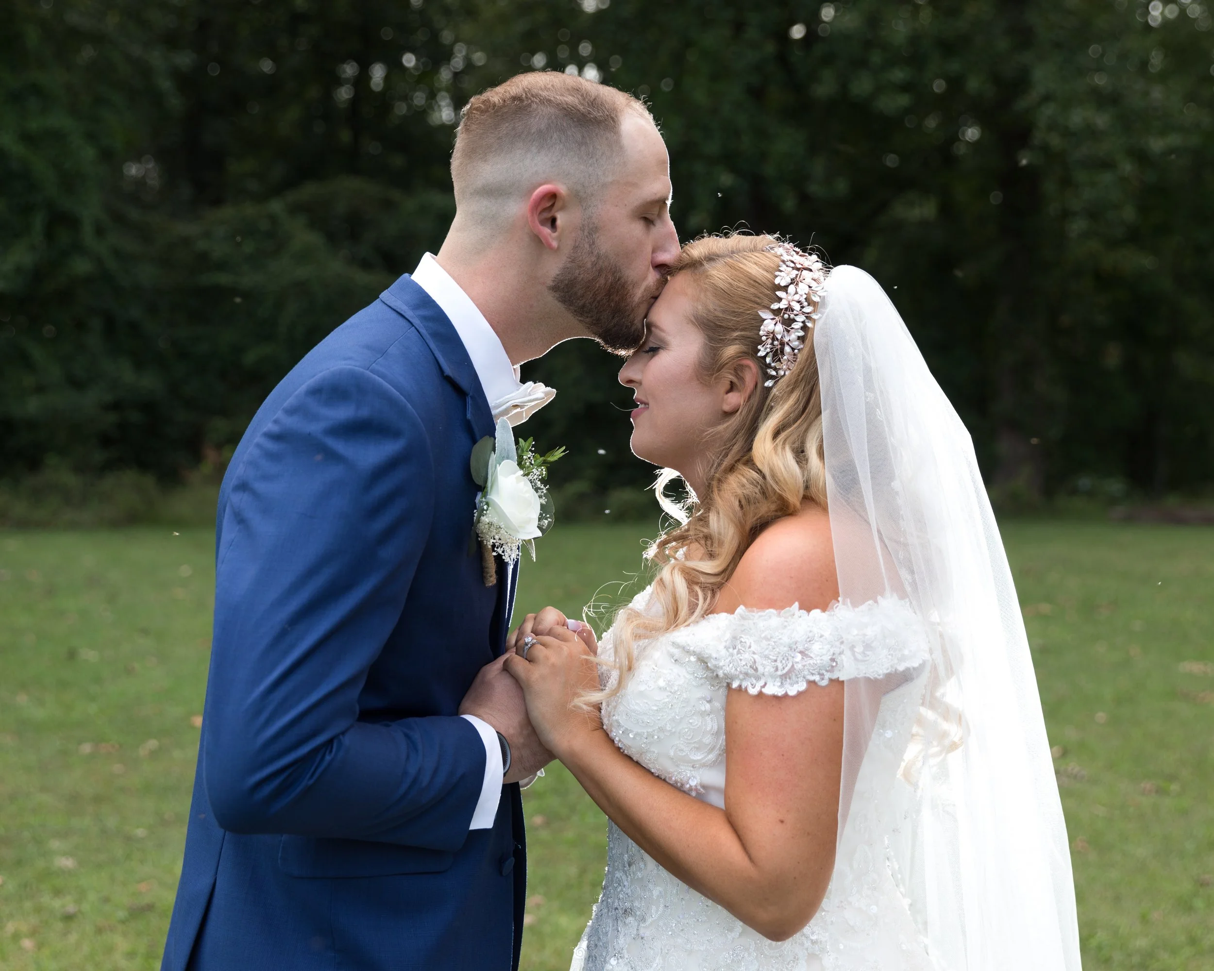Bride and groom in a wedding attire, groom kissing bride's forehead outdoors on grass.