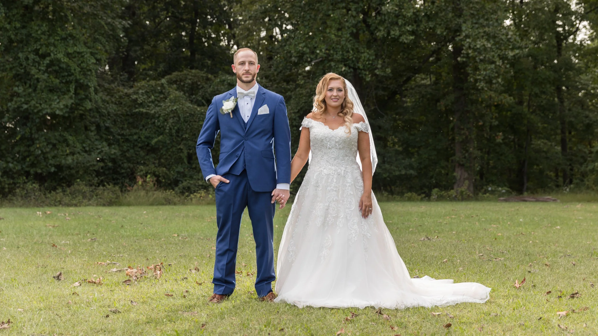 Bride and groom standing outdoors in wedding attire, man in blue suit, woman in white gown, holding hands on grass with trees in background.