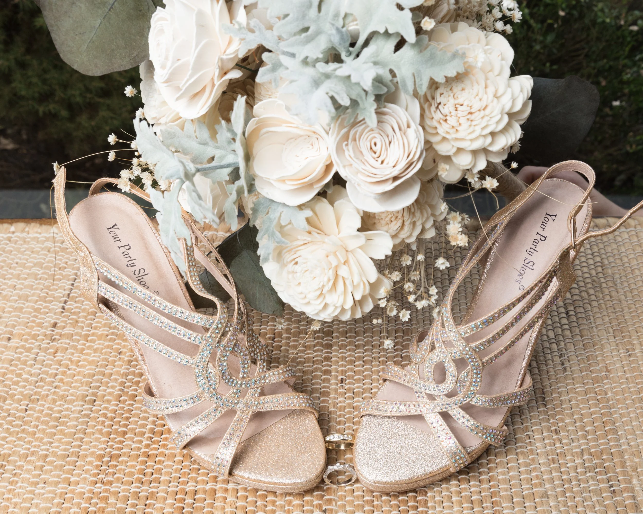 Pair of gold glittery high-heeled sandals with rhinestone embellishments, placed on a woven surface beside a bouquet of cream-colored flowers and dusty miller leaves.