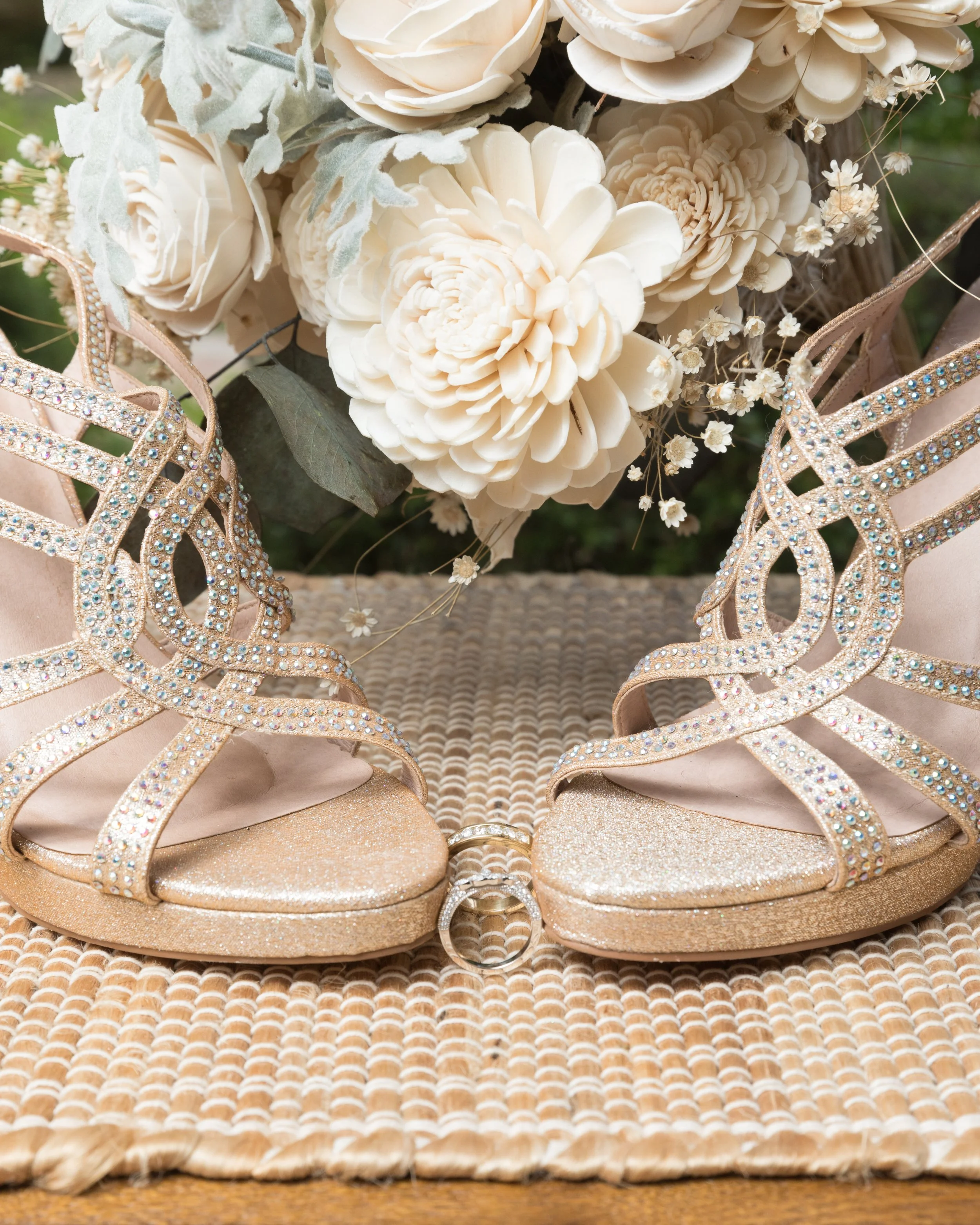 Close-up of sparkling high-heeled shoes with intertwined rings on a woven mat, accompanied by a bouquet of white roses and greenery.