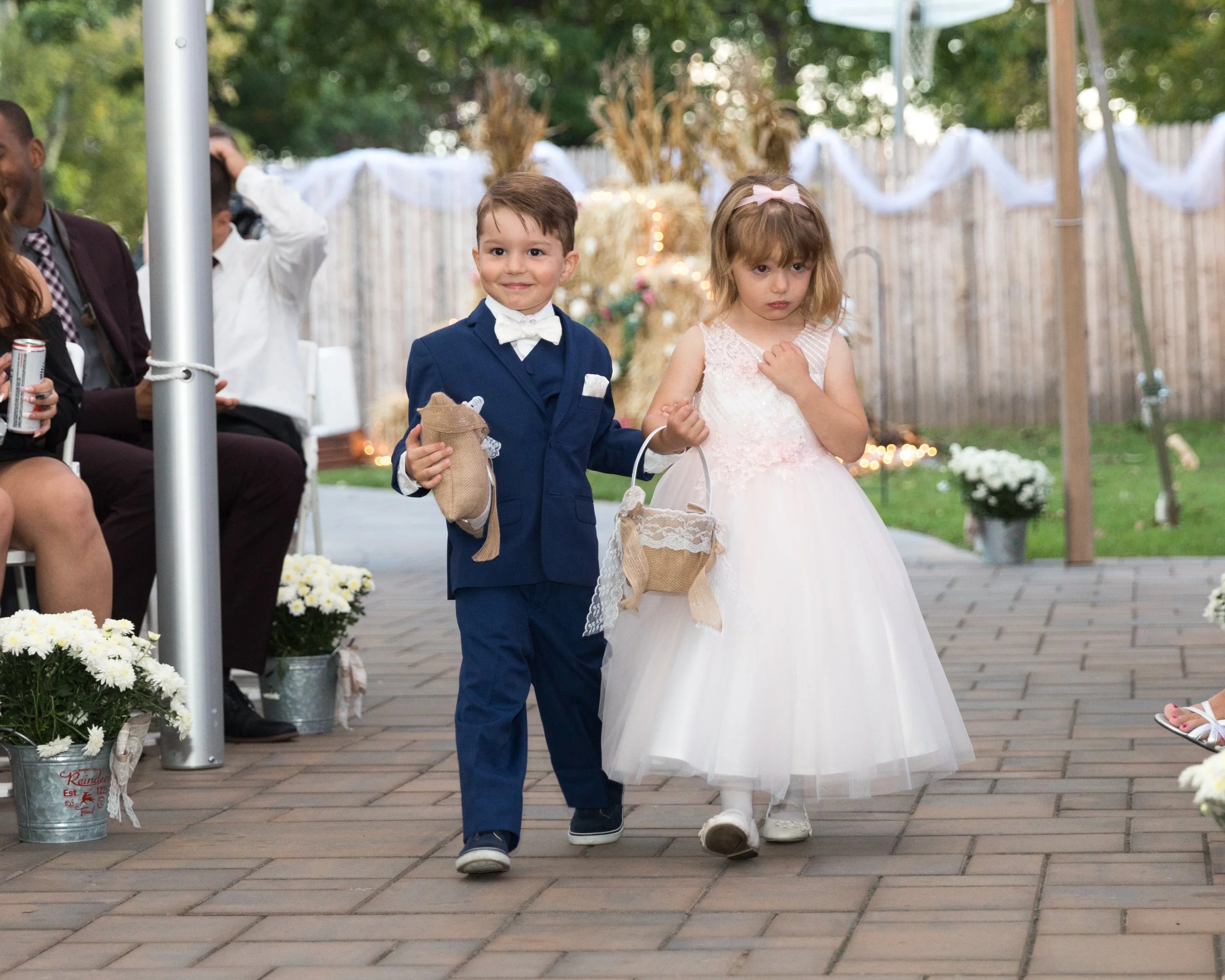 Two children dressed in formal attire walking down an outdoor aisle, with one holding a small pillow and the other holding a basket. Guests are seated on either side, and the area is decorated with flowers and lights.