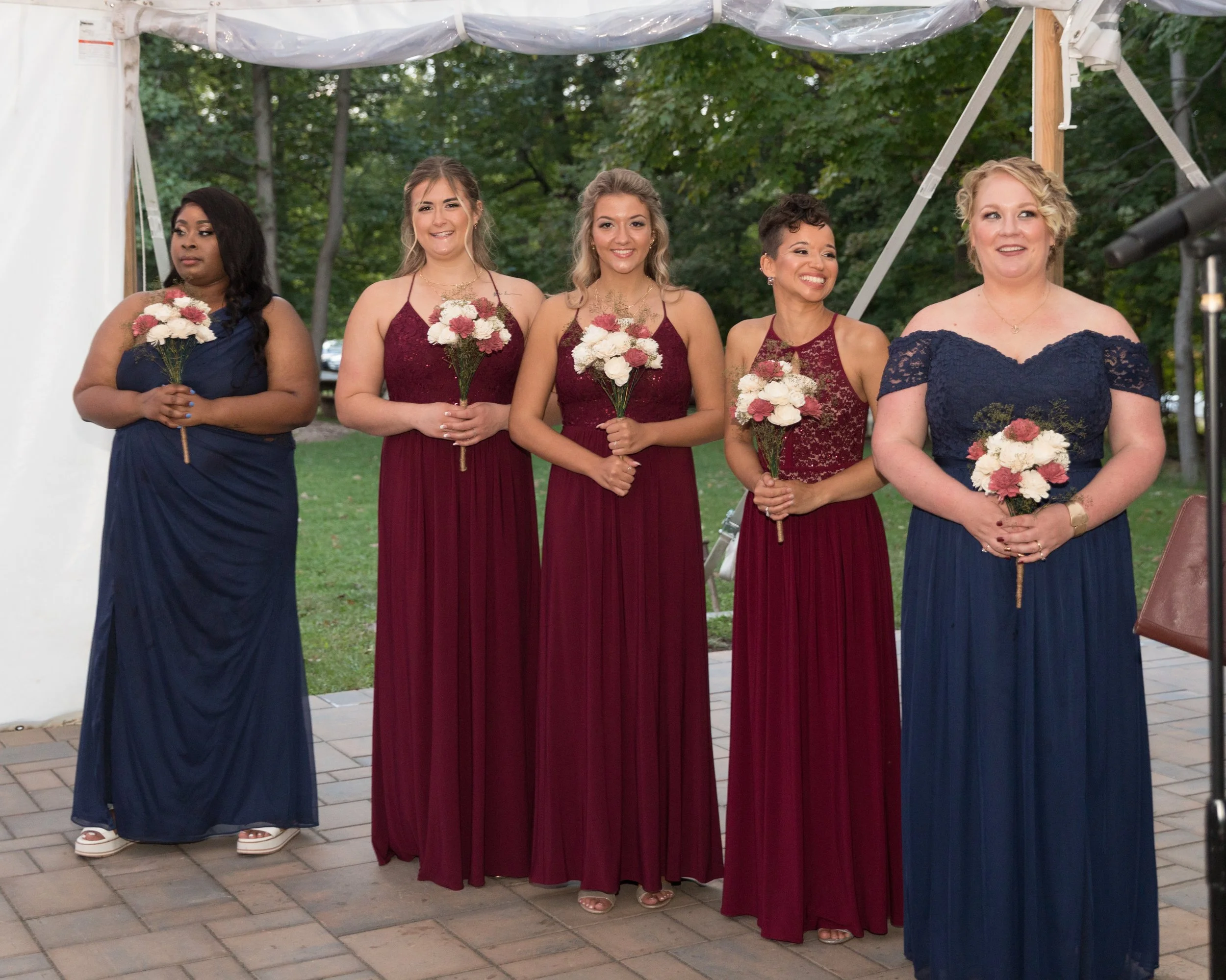 Five women in formal dresses holding bouquets, standing in a tented outdoor setting.