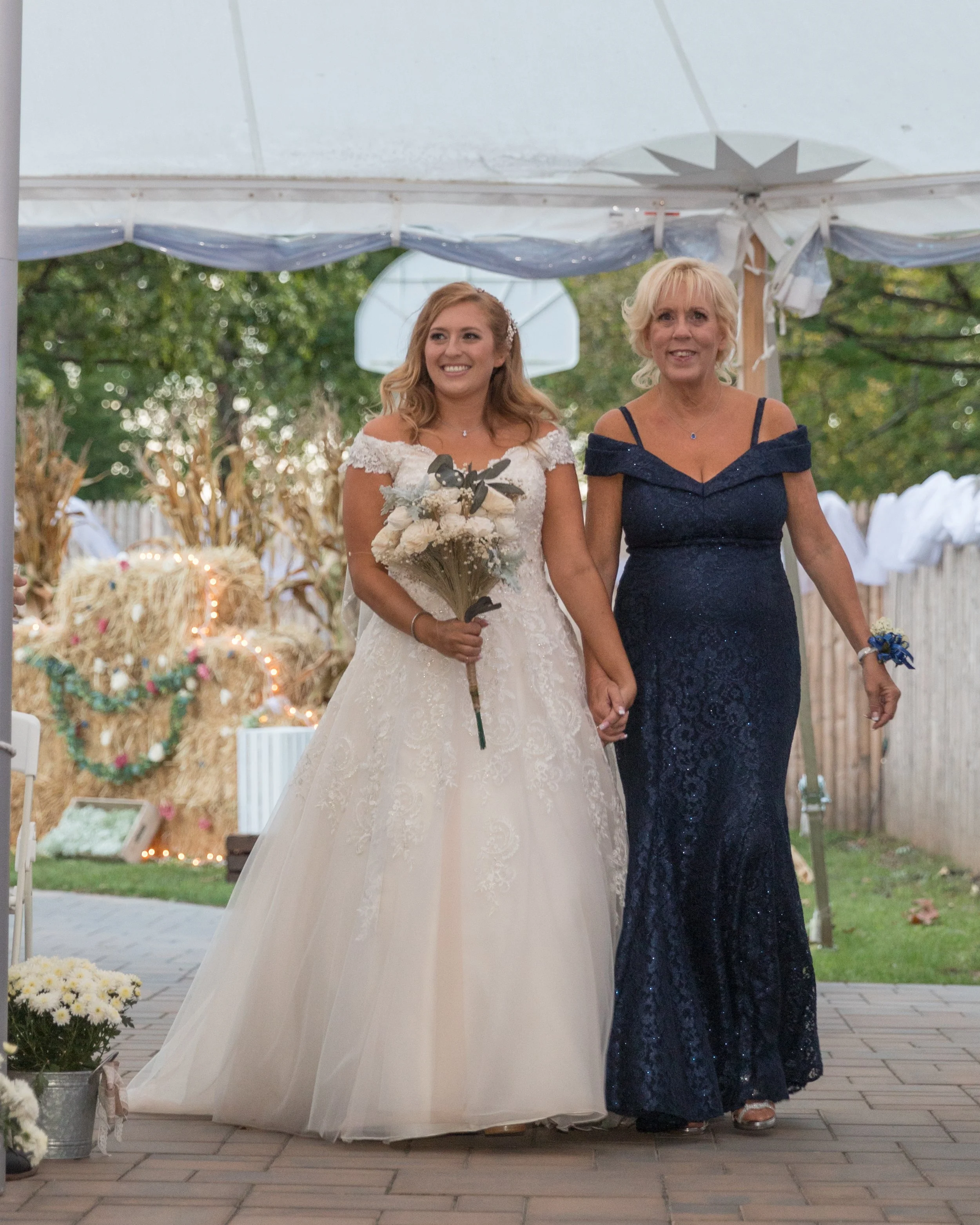 A bride in a white wedding dress holding a bouquet walks hand in hand with a woman in a navy blue gown, likely during a wedding ceremony under a tent with fall-themed decorations.