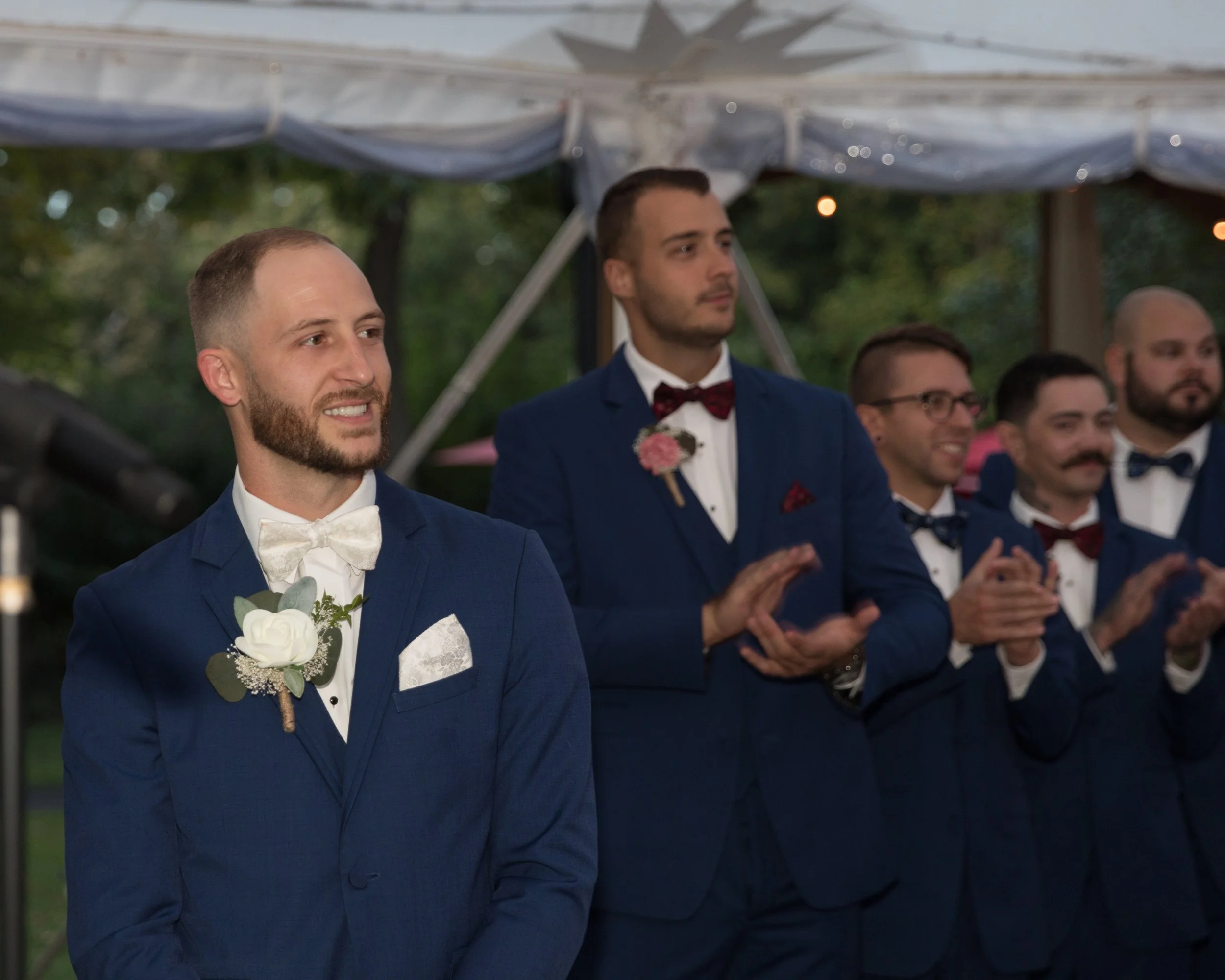 Groom and groomsmen in blue suits clapping at a wedding ceremony.