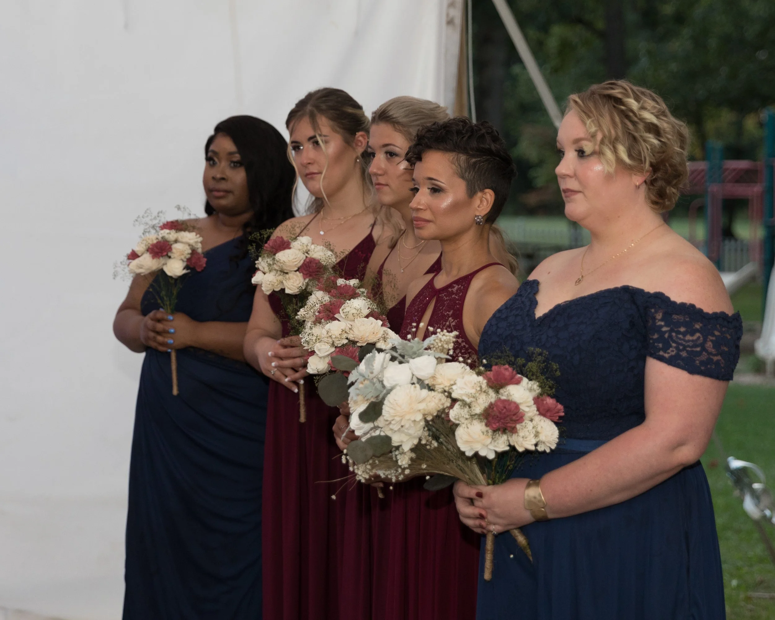 A group of bridesmaids in burgundy and navy dresses holding bouquets of flowers, posing together at an outdoor event.