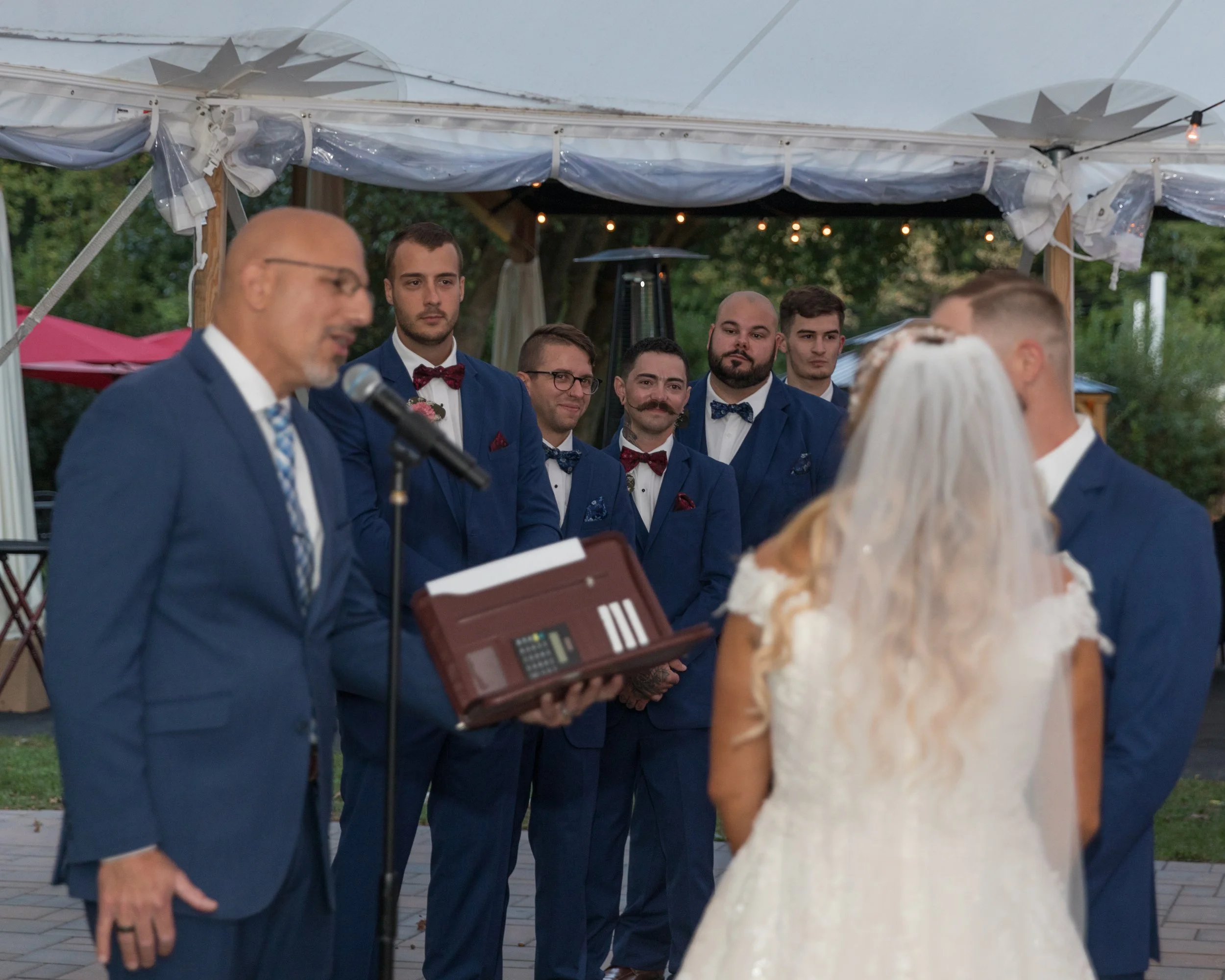 Wedding ceremony outdoors under a tent with a bald man in a suit speaking at a microphone while a groom and bride face each other. Groomsmen in blue suits and bow ties stand in the background.