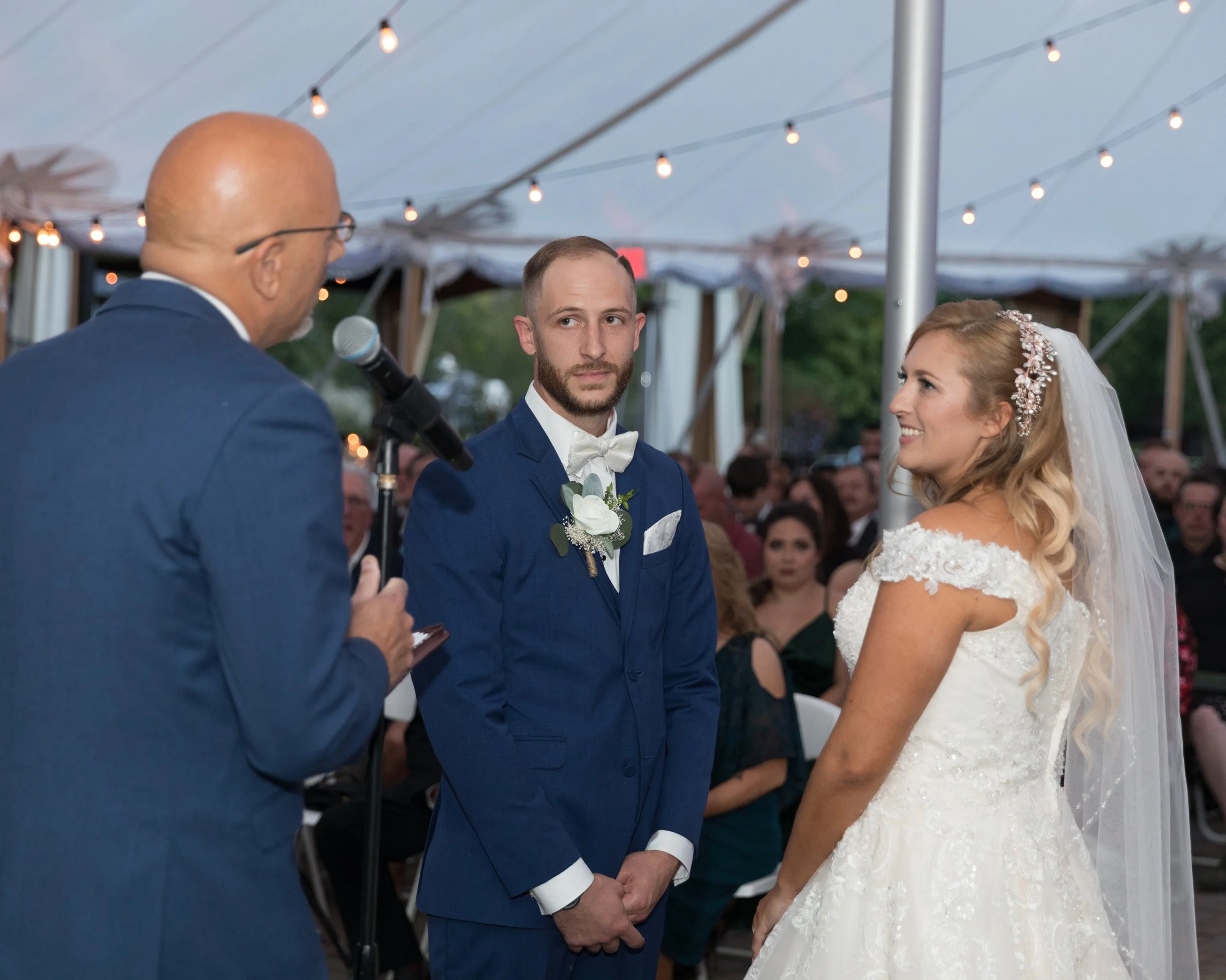 Bride and groom standing under a tent during their wedding ceremony, with an officiant speaking into a microphone.