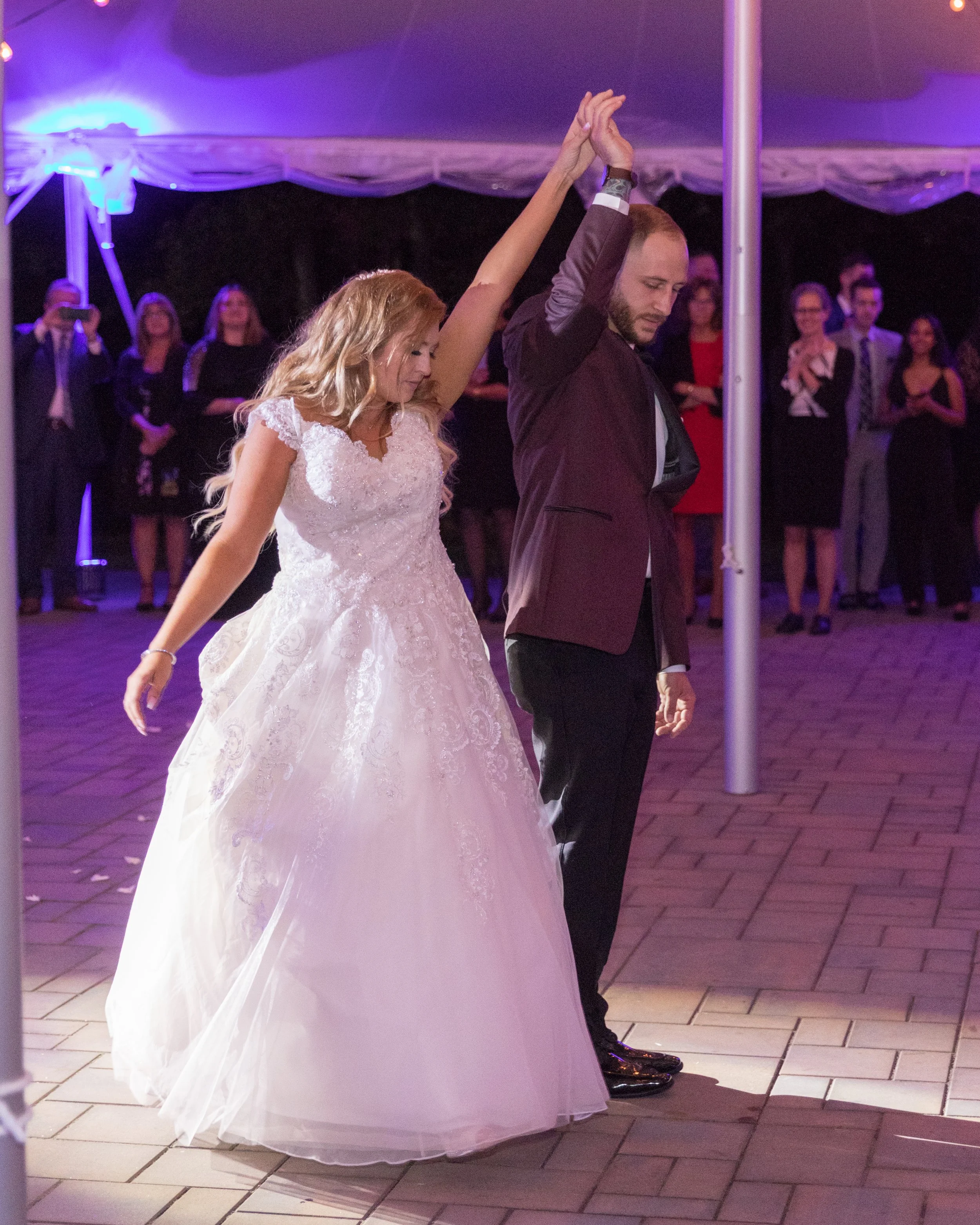 Bride and groom dancing at wedding reception, under purple-lit canopy, surrounded by guests.