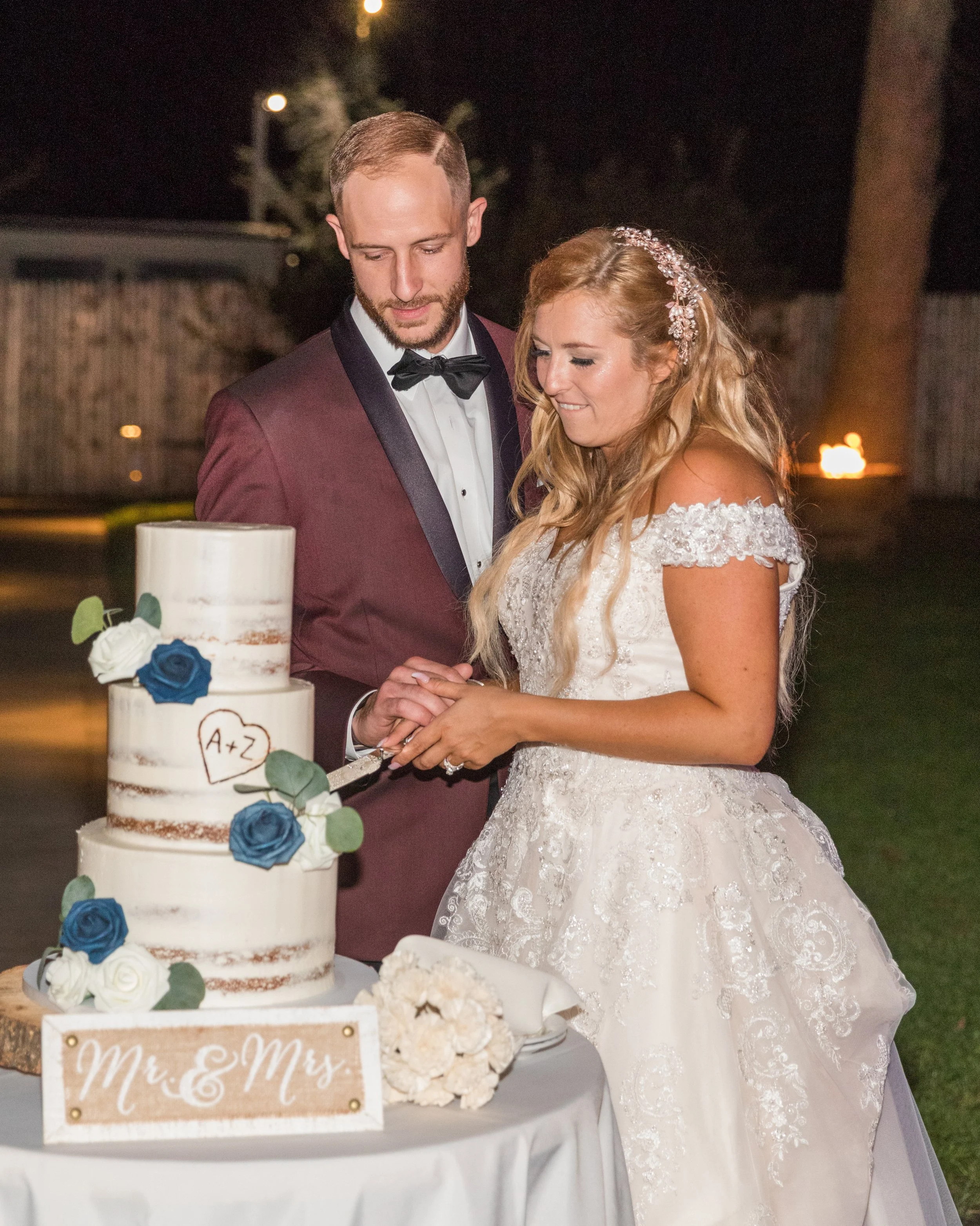 A bride and groom cutting a three-tiered white wedding cake decorated with blue and white flowers and a wooden heart with initials A+Z. They stand at an outdoor venue at night, with a sign reading "Mr & Mrs" on the table.